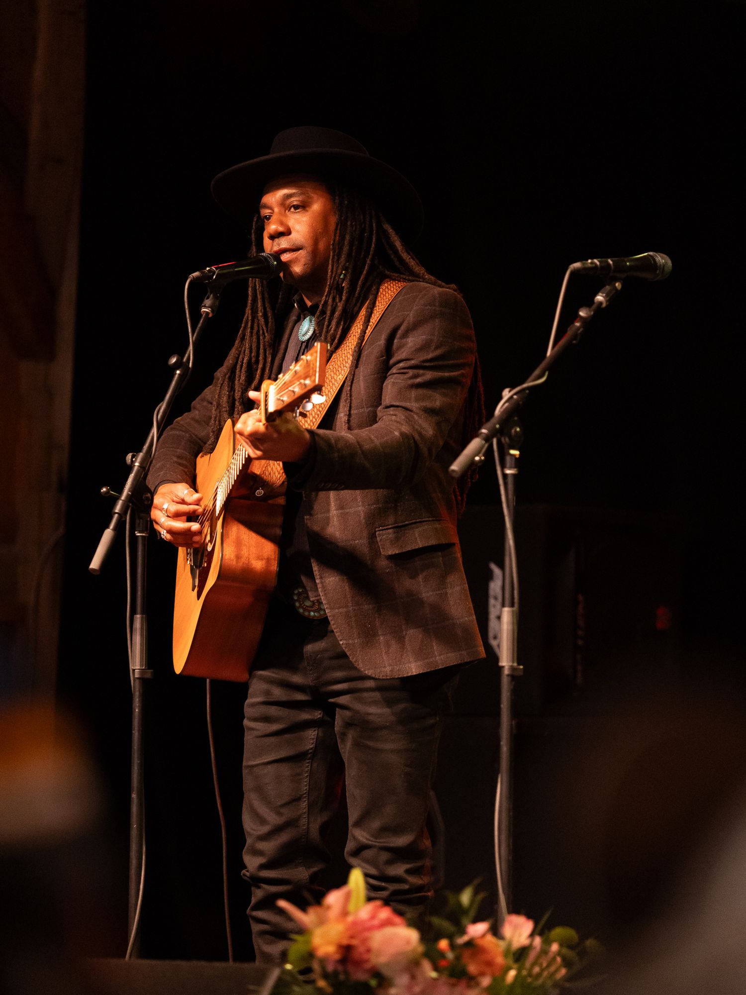 A musician with dreadlocks and a black hat plays acoustic guitar on a dimly lit stage.