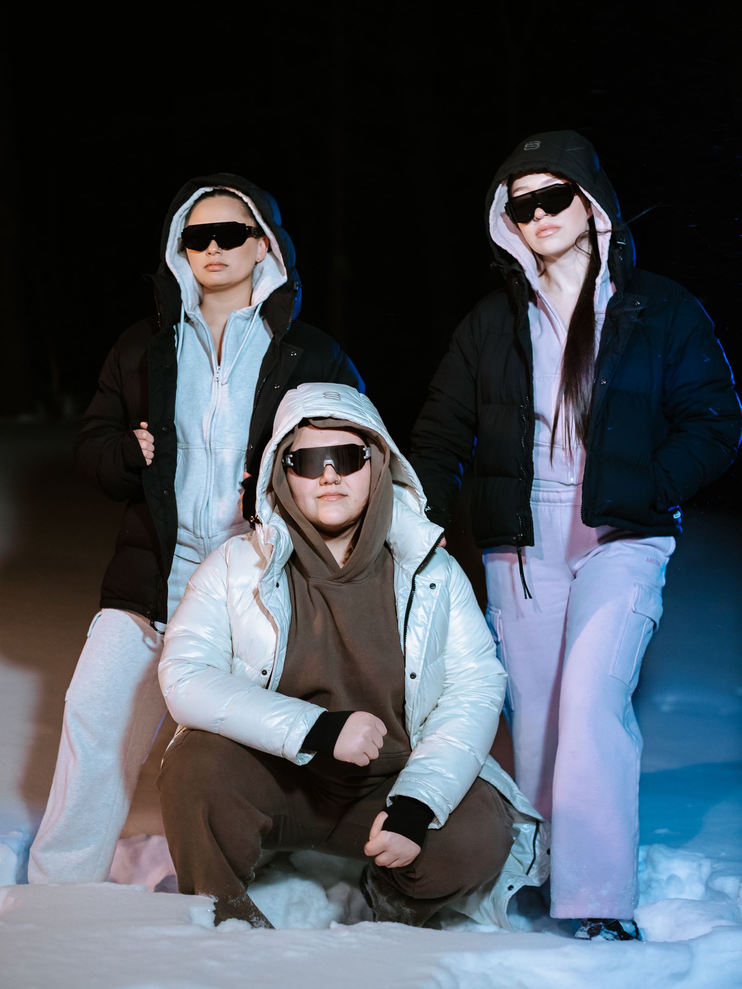 Three models in winter puffer jackets and sunglasses pose dramatically in snow against a dark night background.