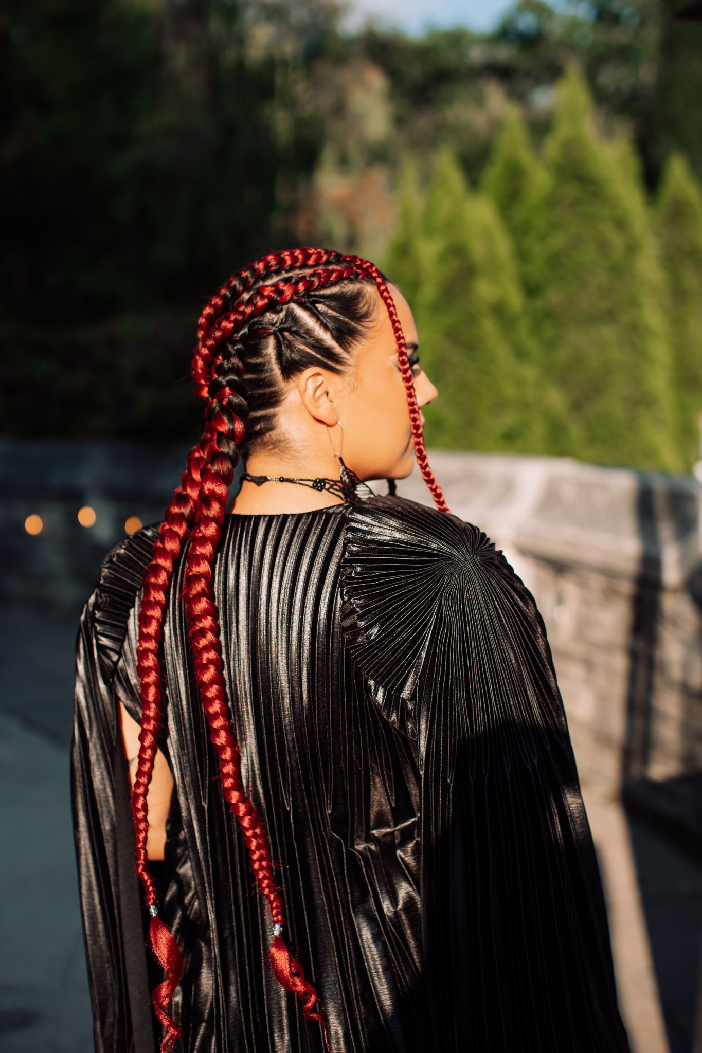 Woman with vibrant red braids wearing a black pleated cape gown at an outdoor fashion event.