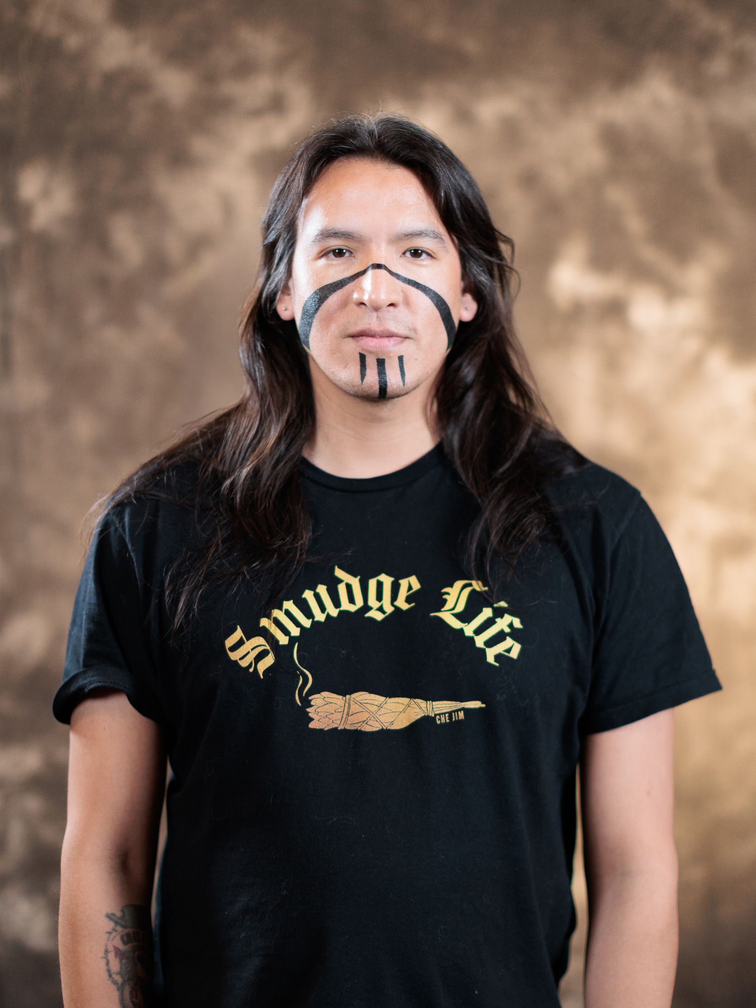 Person with long dark hair and traditional face paint wearing a black Smudge Life t-shirt against a brown backdrop.