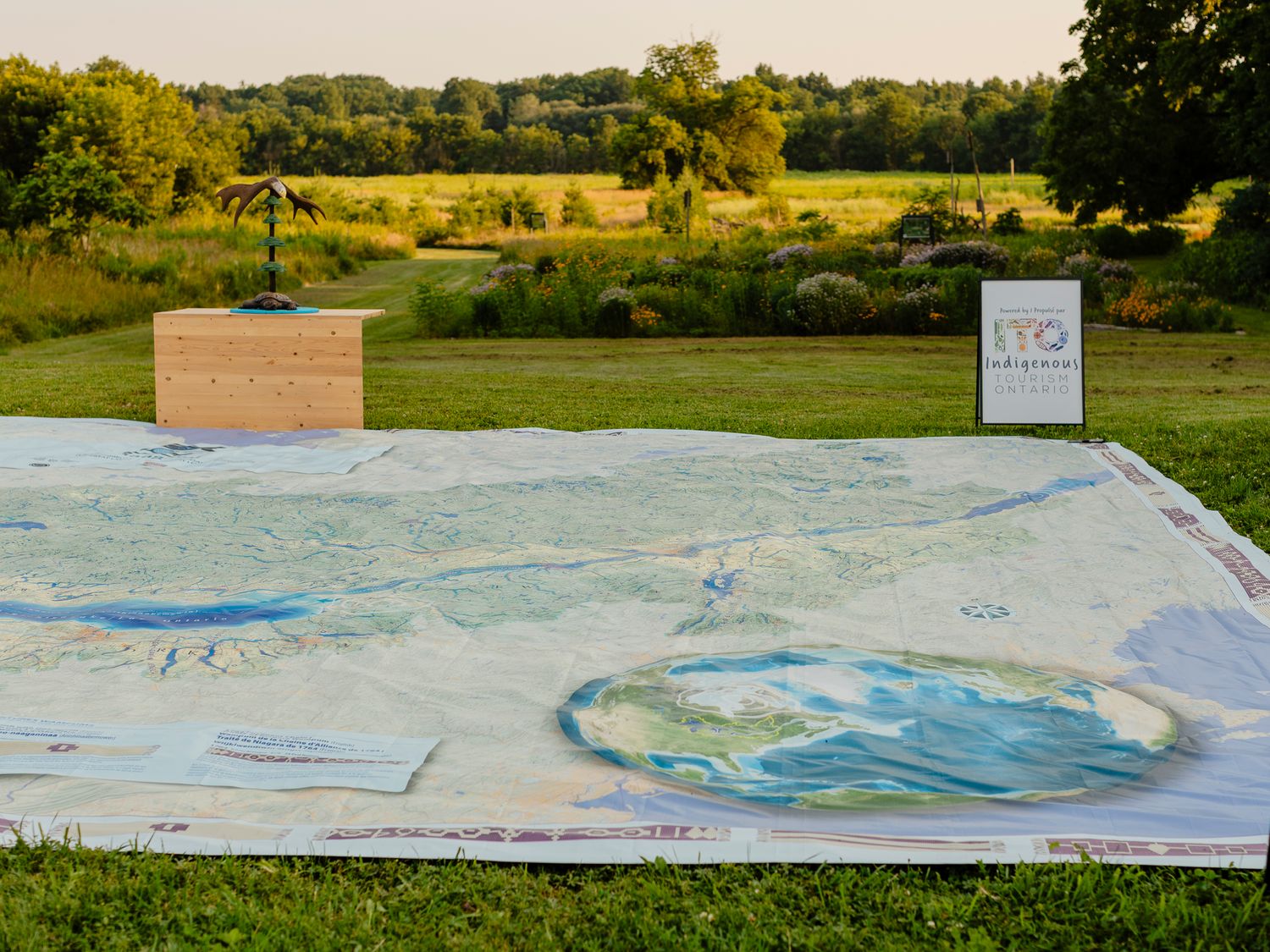 Large outdoor map layout on grass with a wooden display box and informational sign in a scenic green landscape.