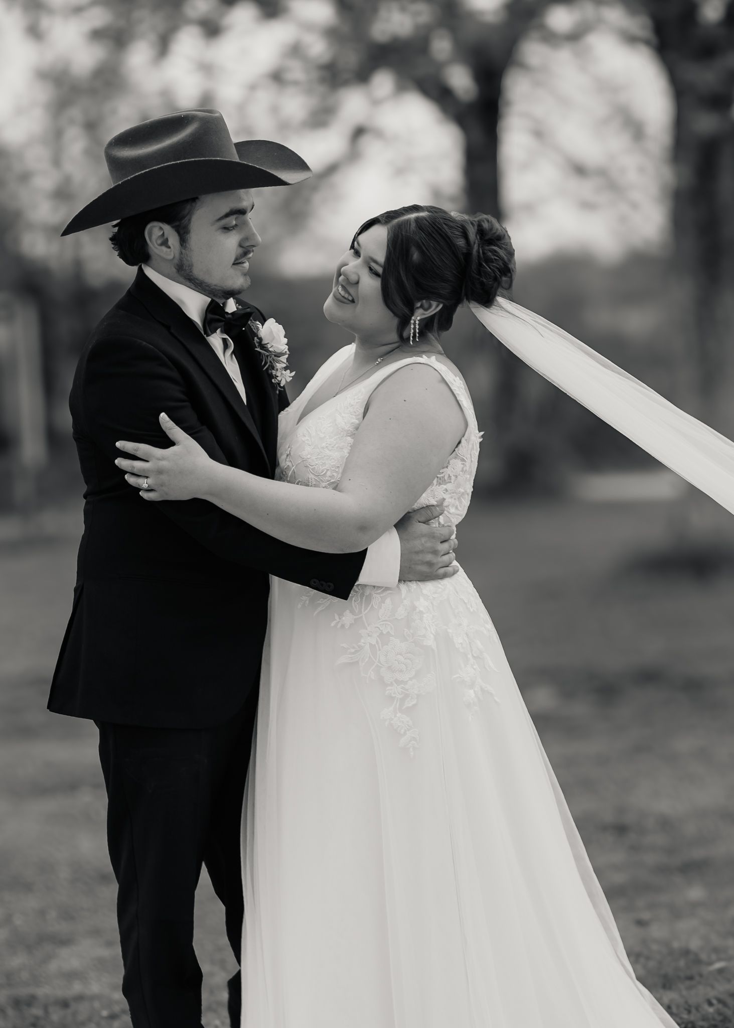Black and white portrait of bride and groom embracing outdoors with veil flowing in wind.