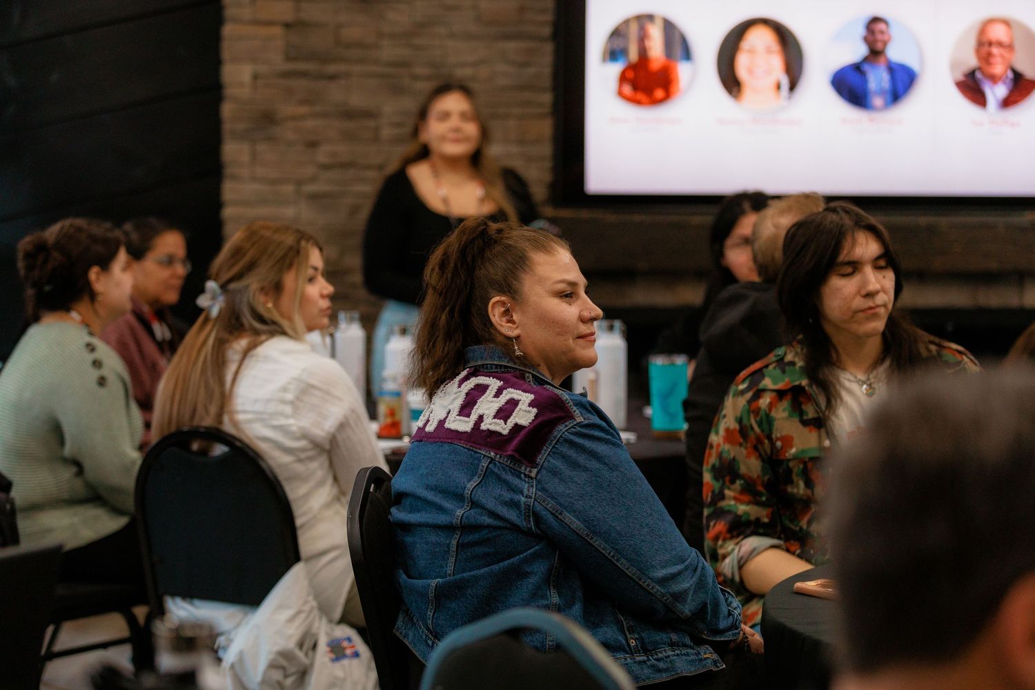 Diverse group of women attending a community seminar, listening attentively with a presentation screen visible in background.