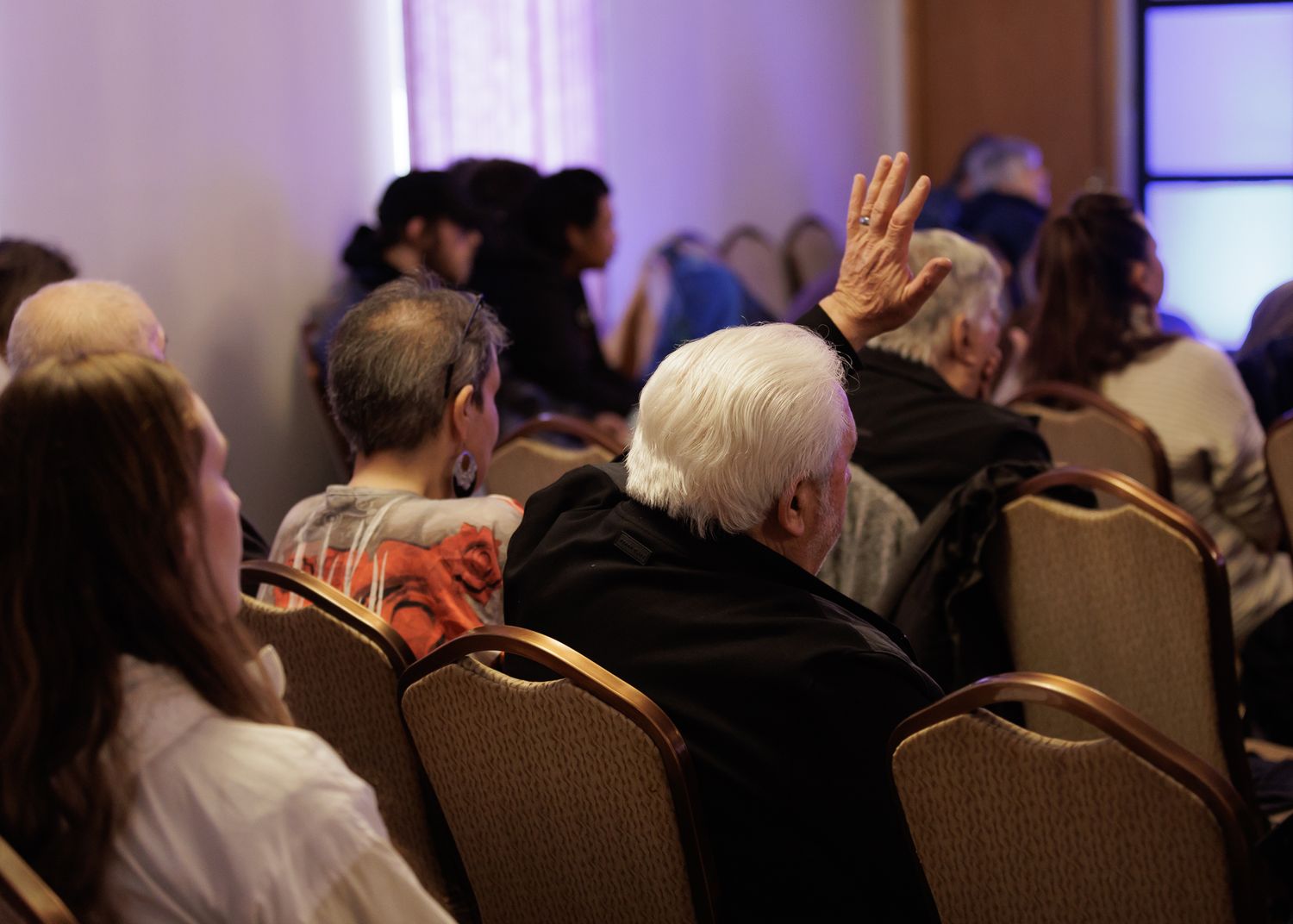 Audience members seated in chairs at a conference, with an elderly person raising their hand during a presentation.