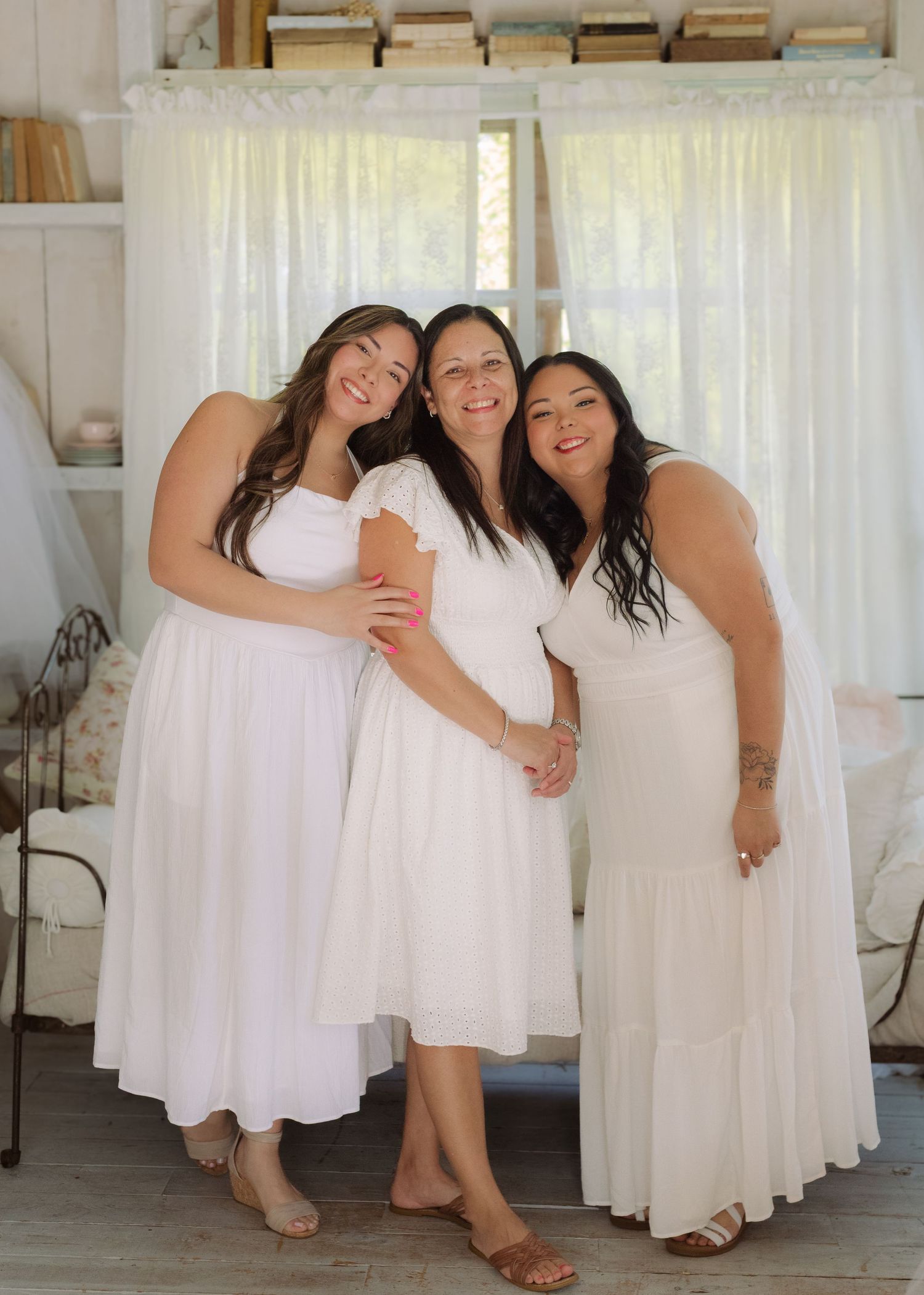 Three women in white dresses smile together indoors in a bright, cozy cottage-style room with sheer curtains.