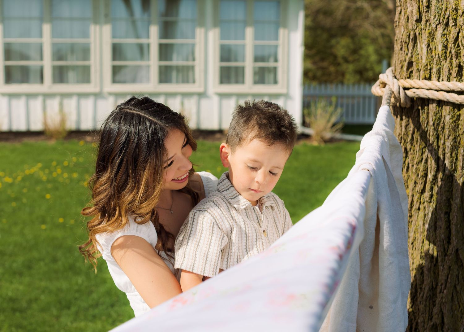 A mother and young boy look at a colorful blanket tied to a tree in a sunny backyard.