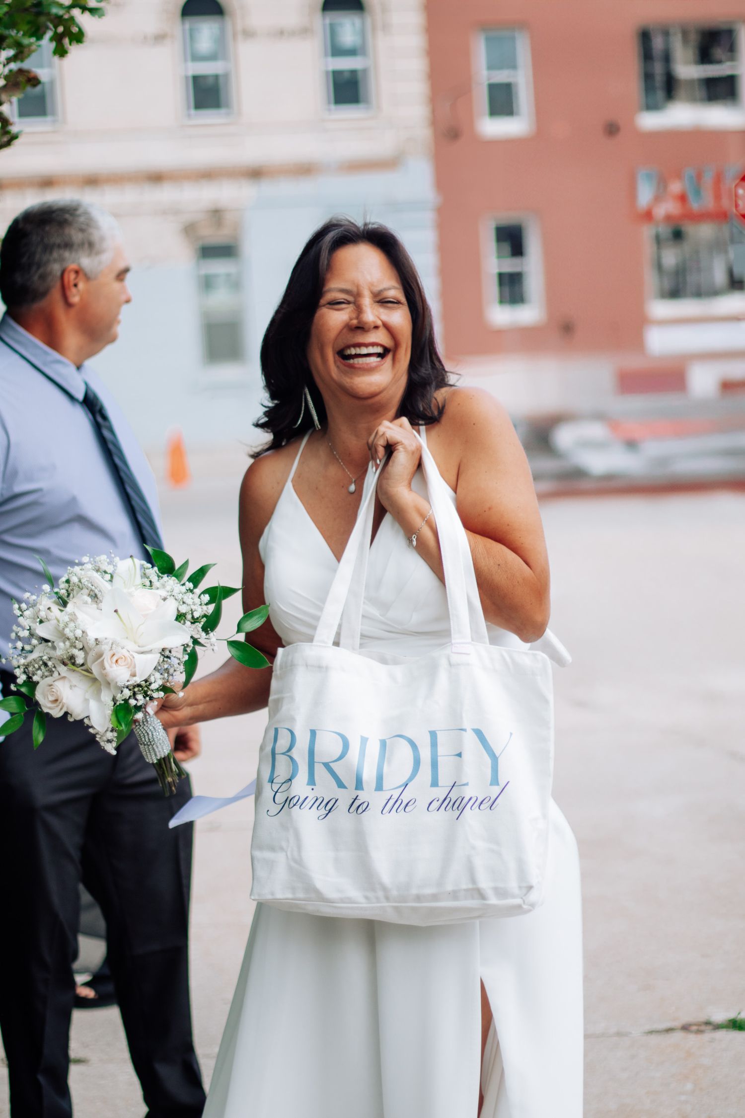 Happy bride laughing outdoors holding white bouquet and Bridey tote bag on wedding day.