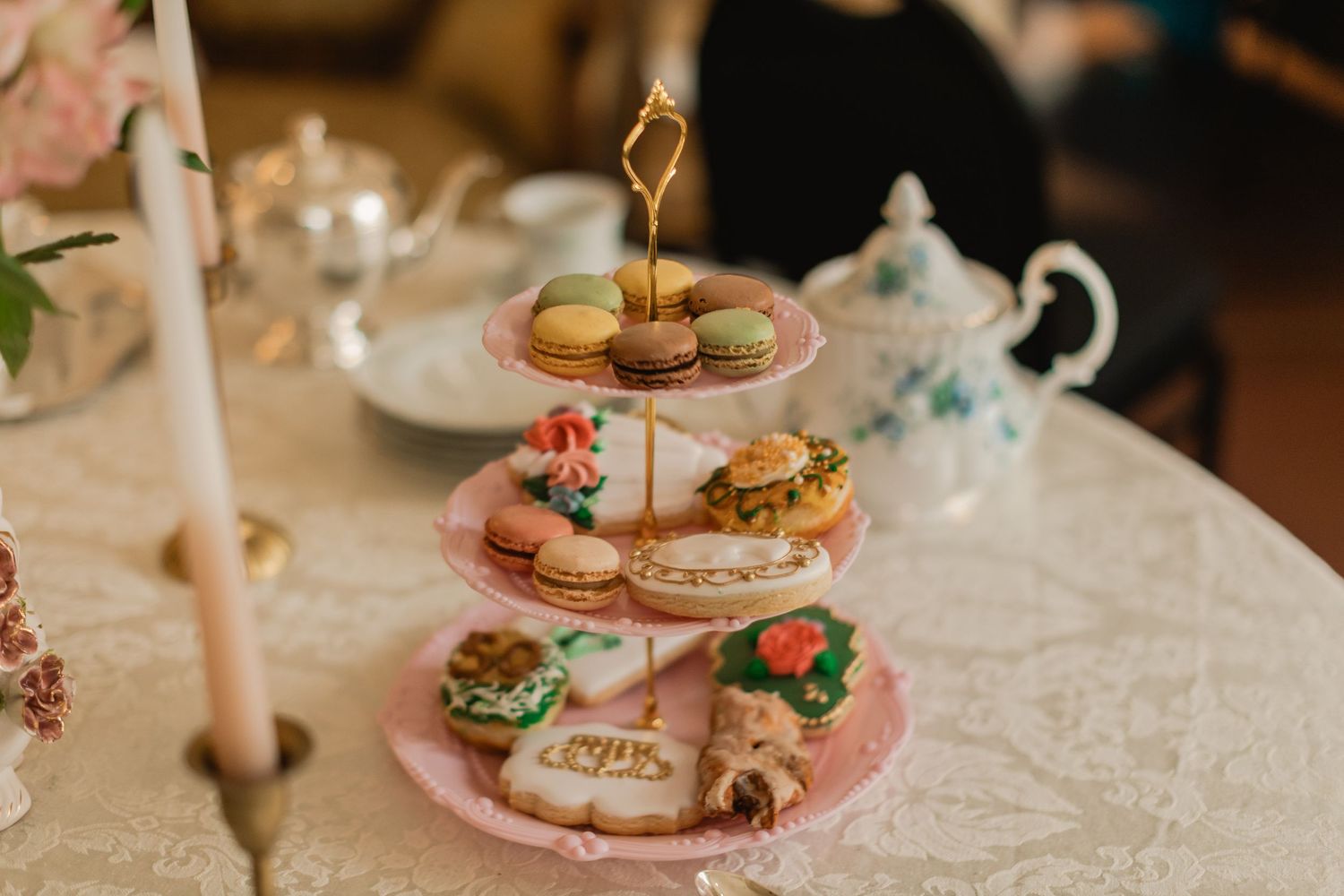 Three-tier pink stand with macarons, sandwiches, and decorated cookies on lace tablecloth at elegant afternoon tea.