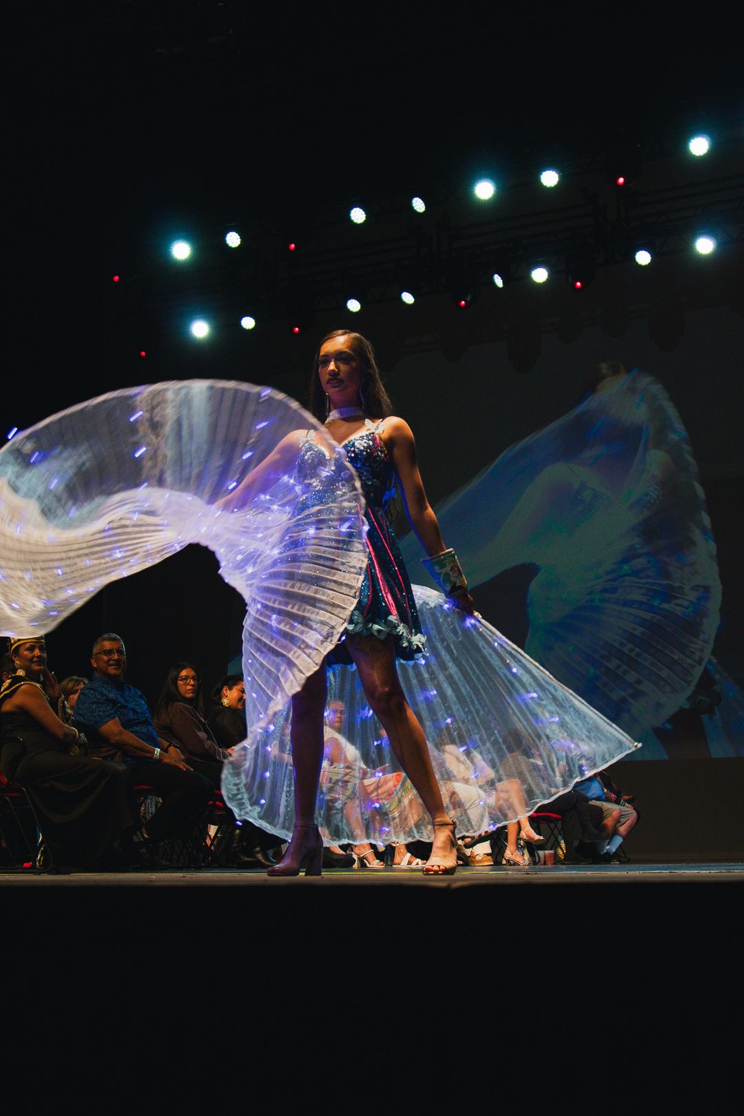 Model walks runway in illuminated butterfly wing dress under stage lights at fashion show.