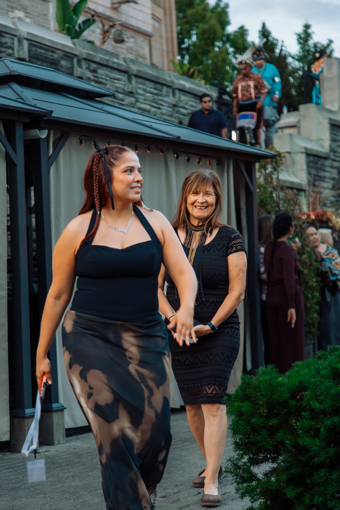Two women walking outdoors, one in black halter top with tie-dye skirt, another in black dress.