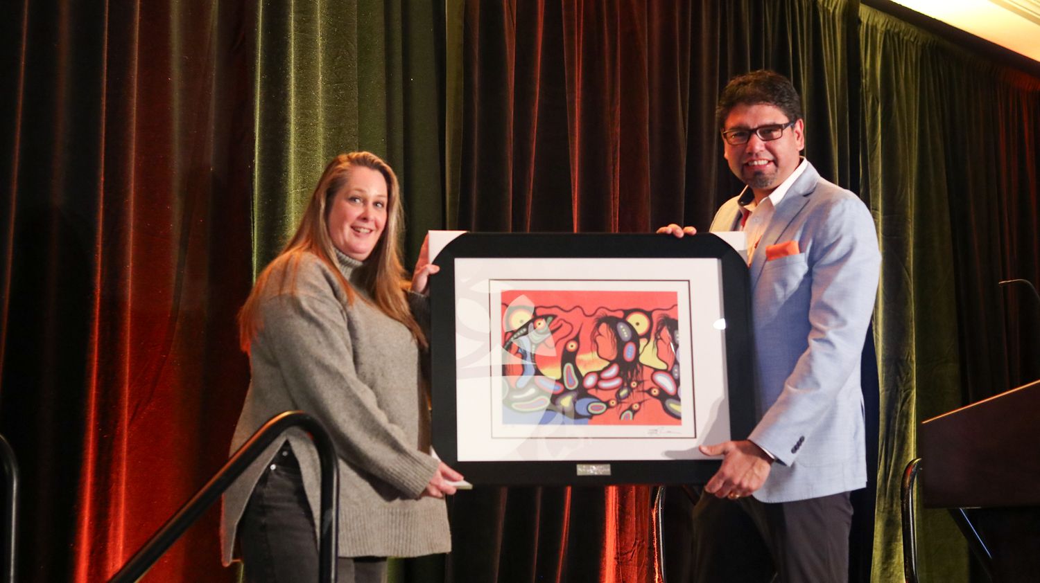 Two people smile while holding a framed colorful artwork at an awards ceremony on stage.