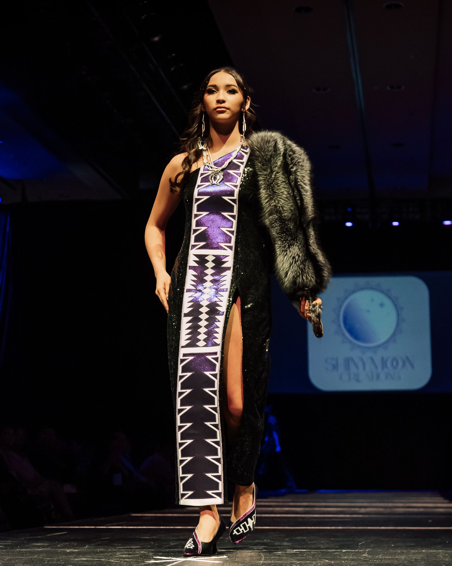 A model walks the runway in a geometric patterned dress with a fur shawl.