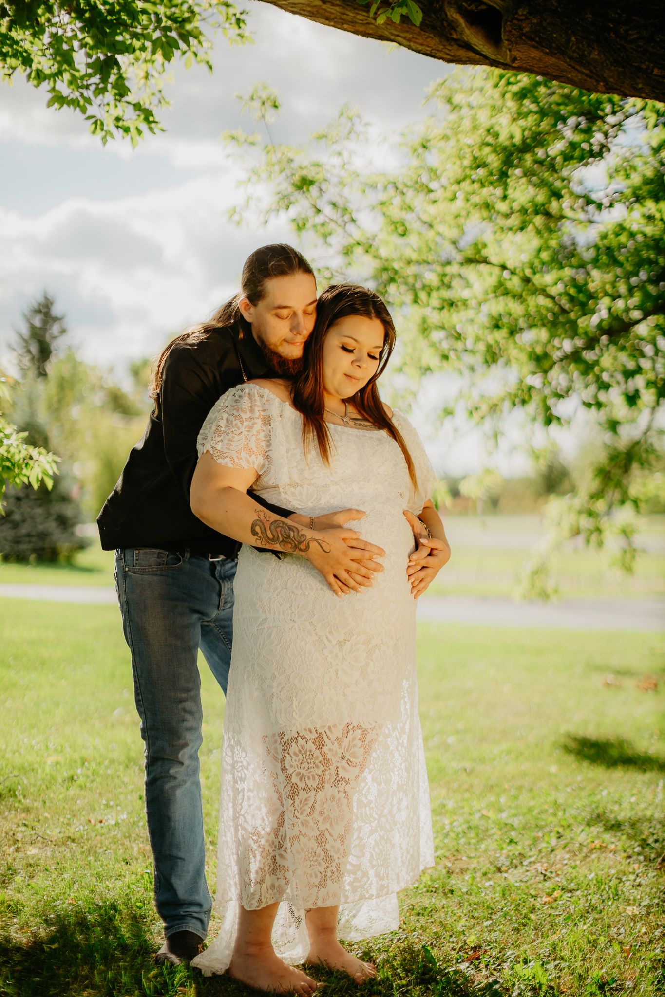 A tender maternity photoshoot moment under a tree in golden sunlight.