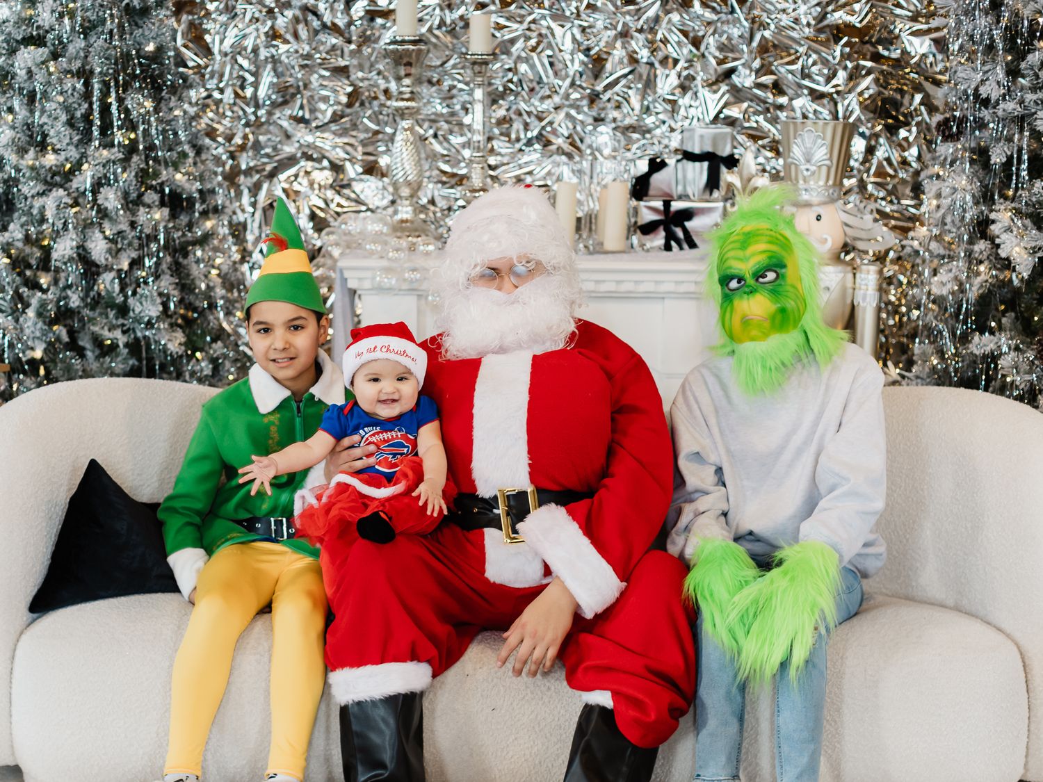 Holiday photo featuring festive characters including Santa Claus, an elf, and the Grinch sitting together on a white couch.
