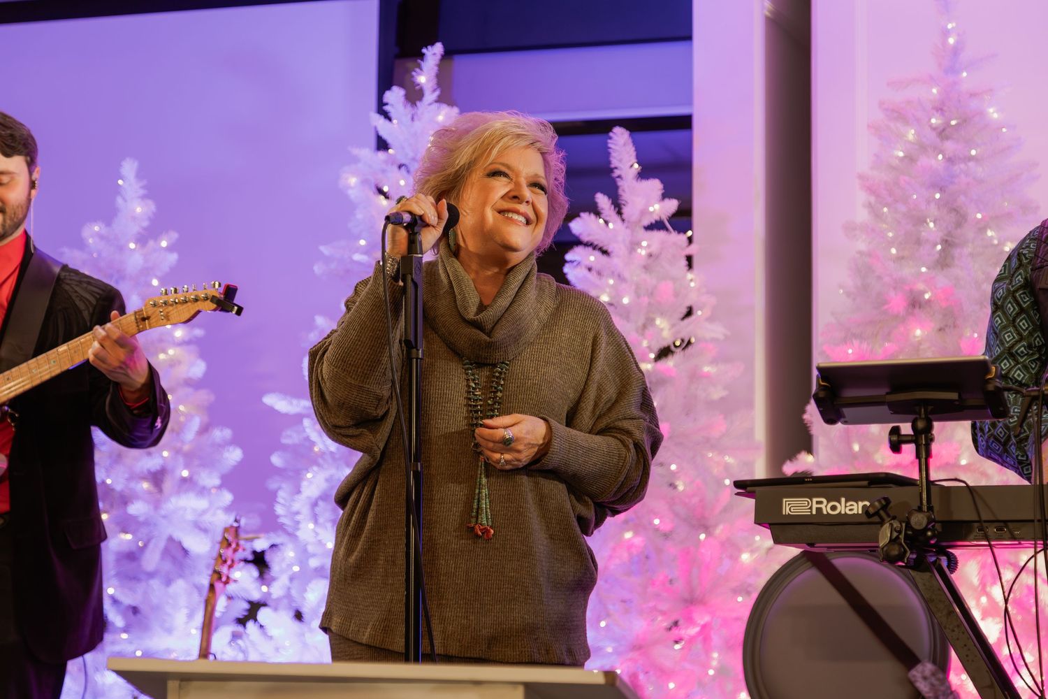 Musicians perform on stage with illuminated white Christmas trees in the background.