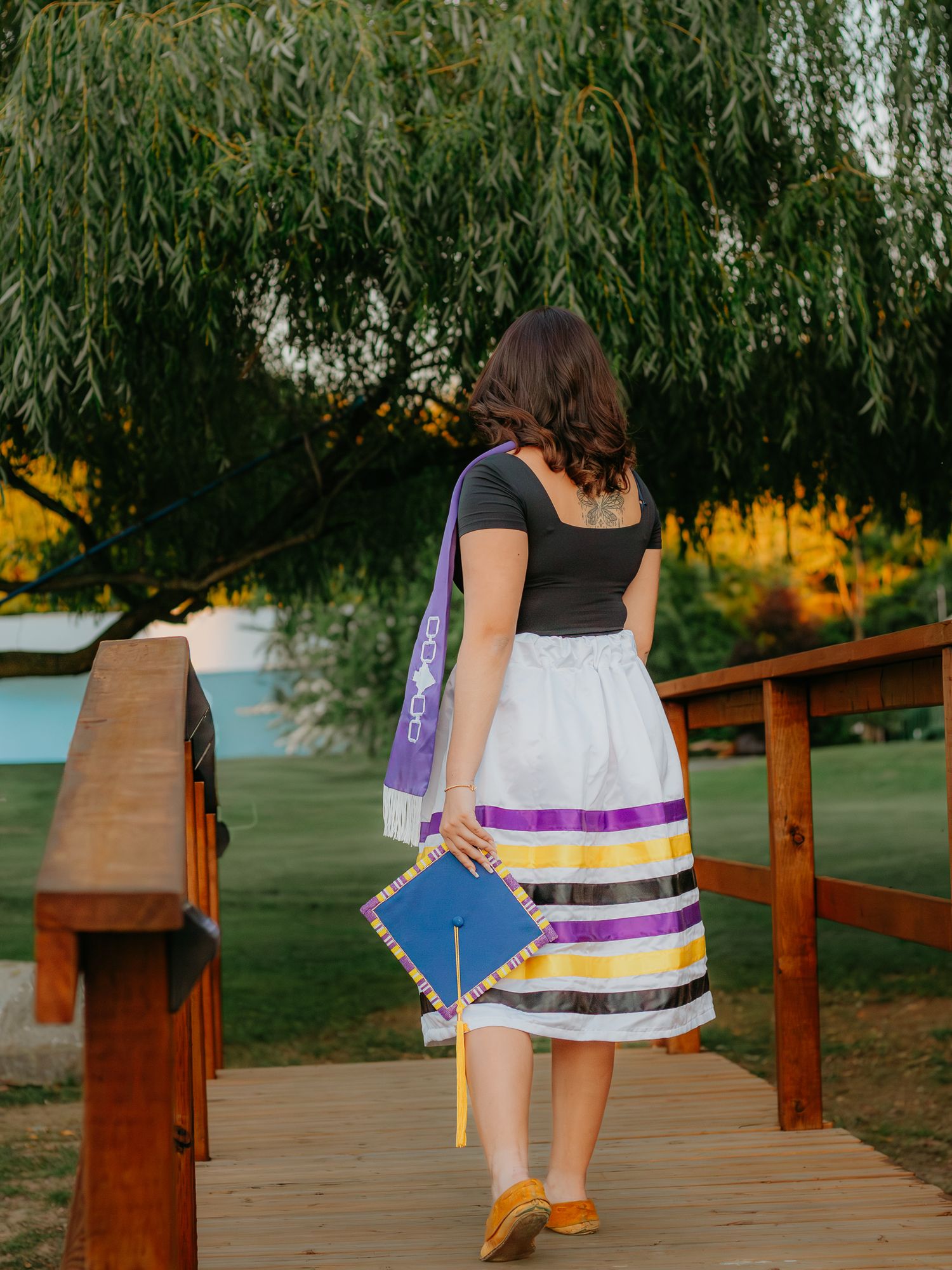 Graduate in striped dress stands on wooden boardwalk under willow trees.