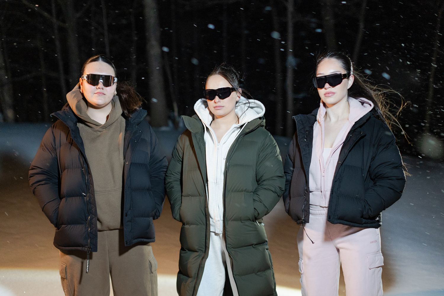 Three people wearing puffer jackets and sunglasses posing together in a snowy nighttime outdoor scene.