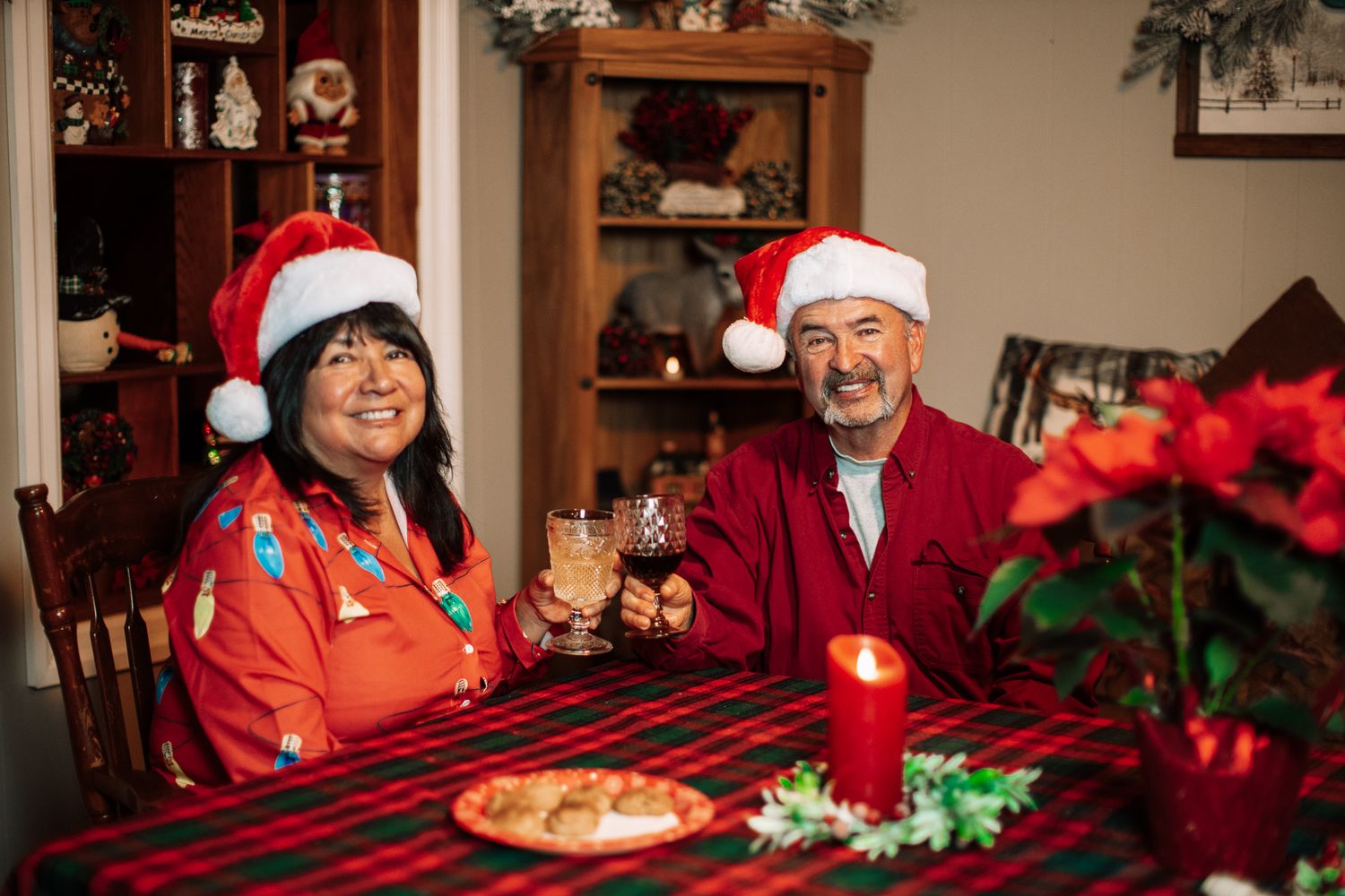 Couple in Santa hats smiling at Christmas dinner table with candle, poinsettia, and holiday decorations.