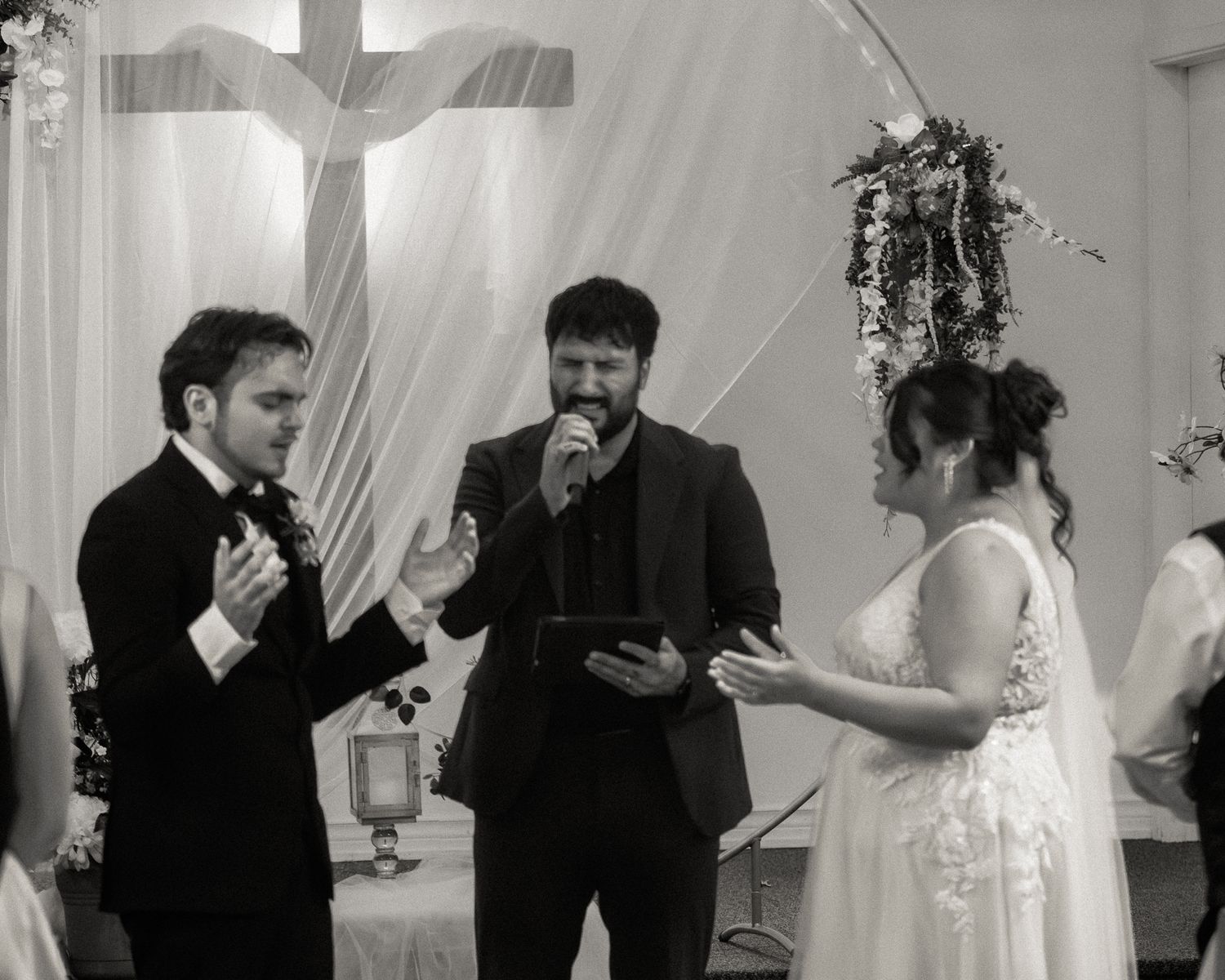 Black and white wedding ceremony photo with bride, groom, and officiant exchanging vows before a cross.