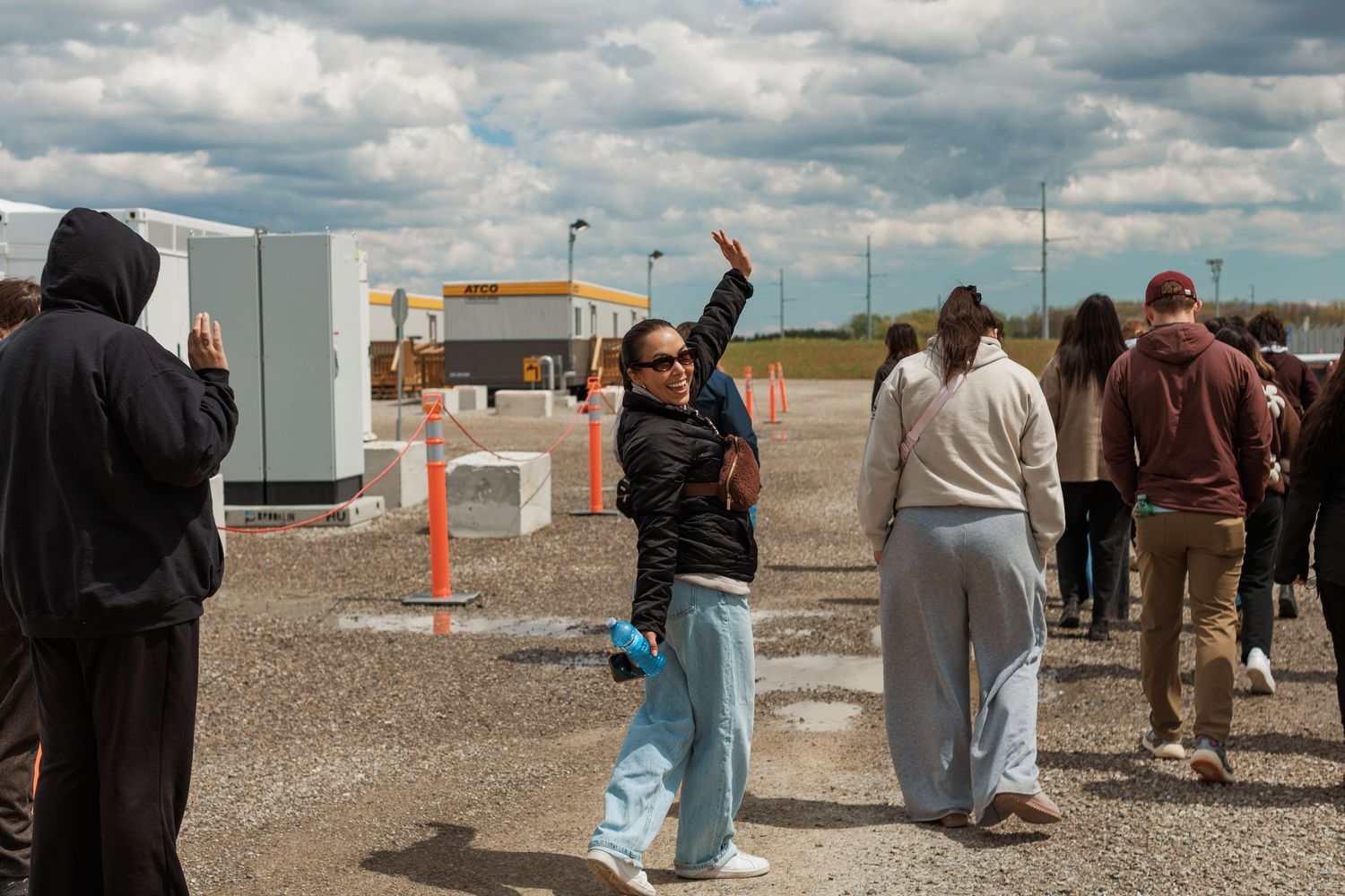 A woman waves while guiding a group of students on an outdoor tour of an industrial facility.