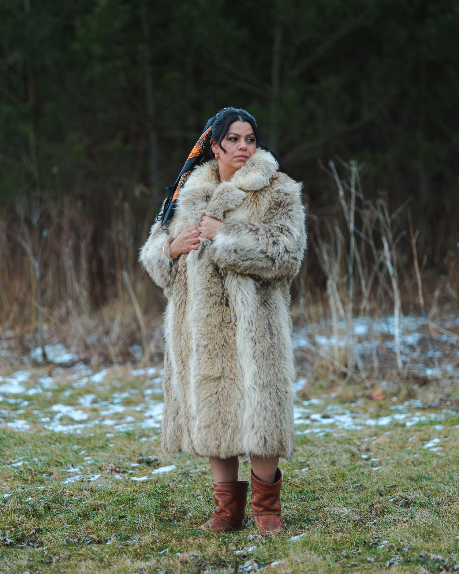 Woman in long fur coat and brown boots stands in snowy field with braided hair accessory.