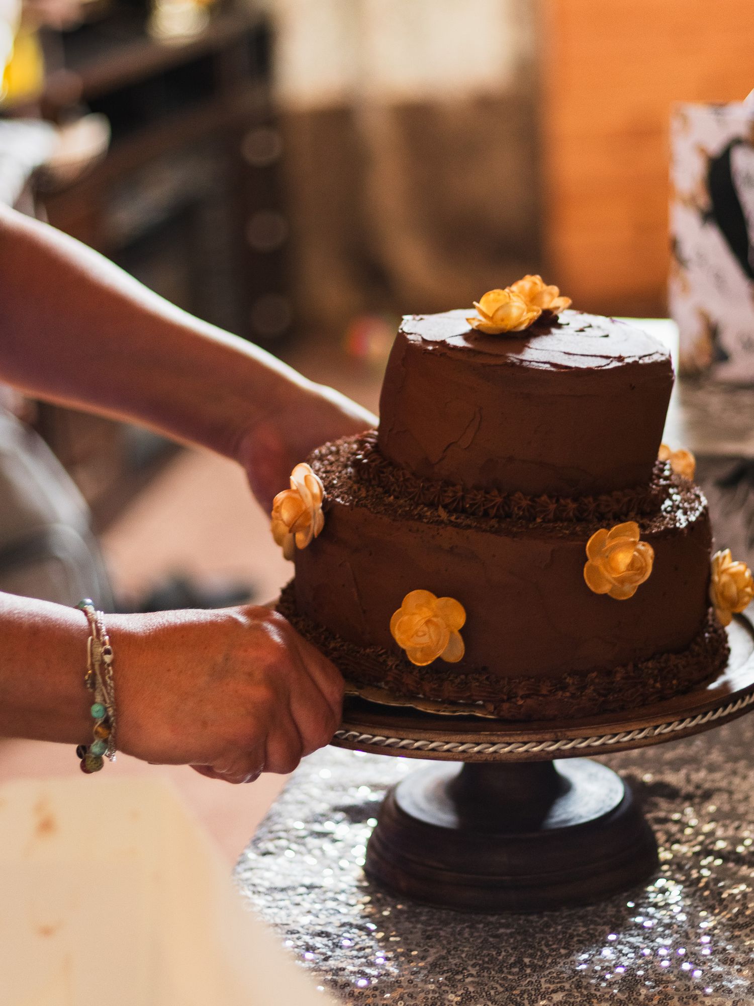 Two-tier chocolate wedding cake decorated with golden flowers being placed on silver cake stand.