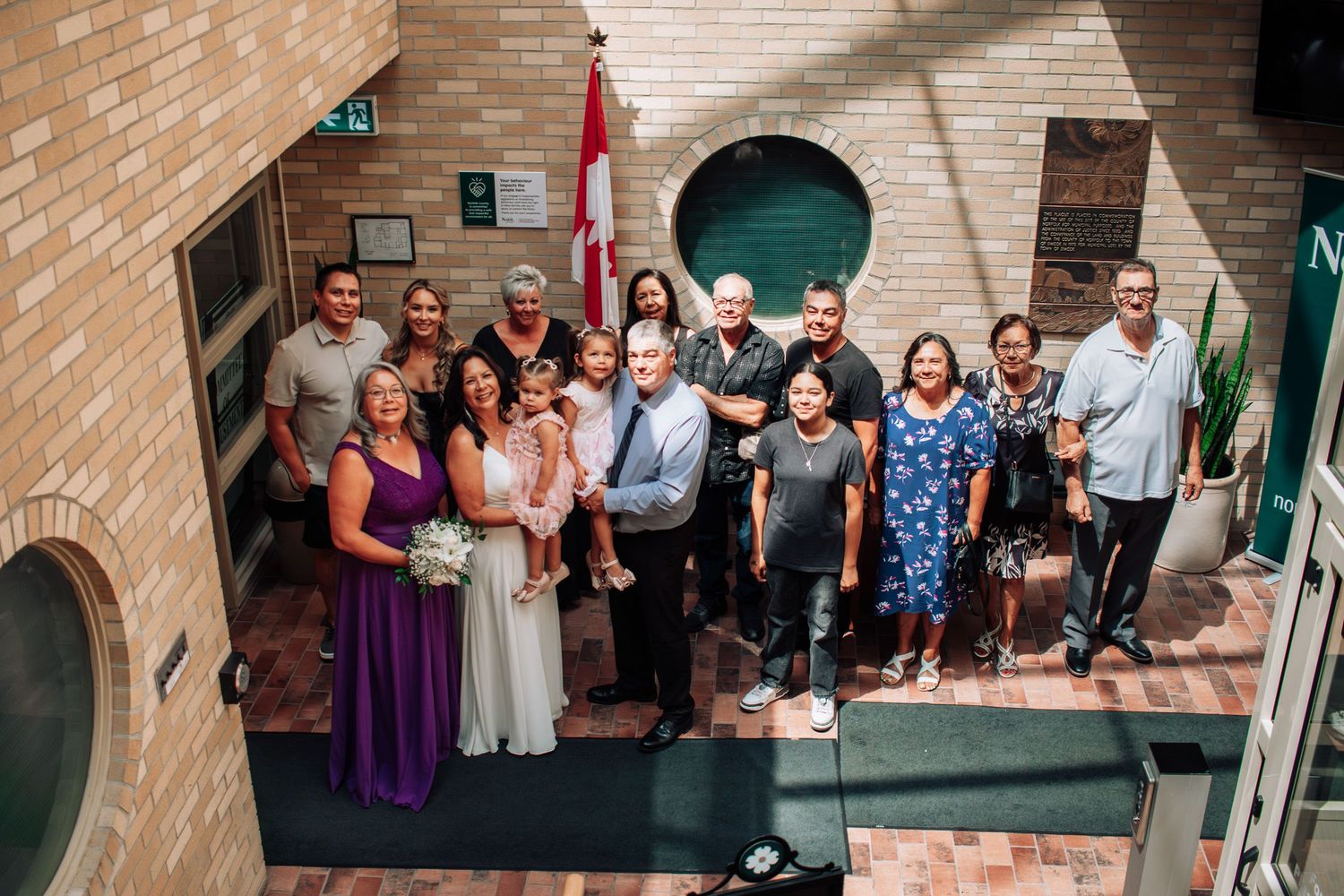 Group of wedding guests posing indoors near a Canadian flag in a brick building atrium.