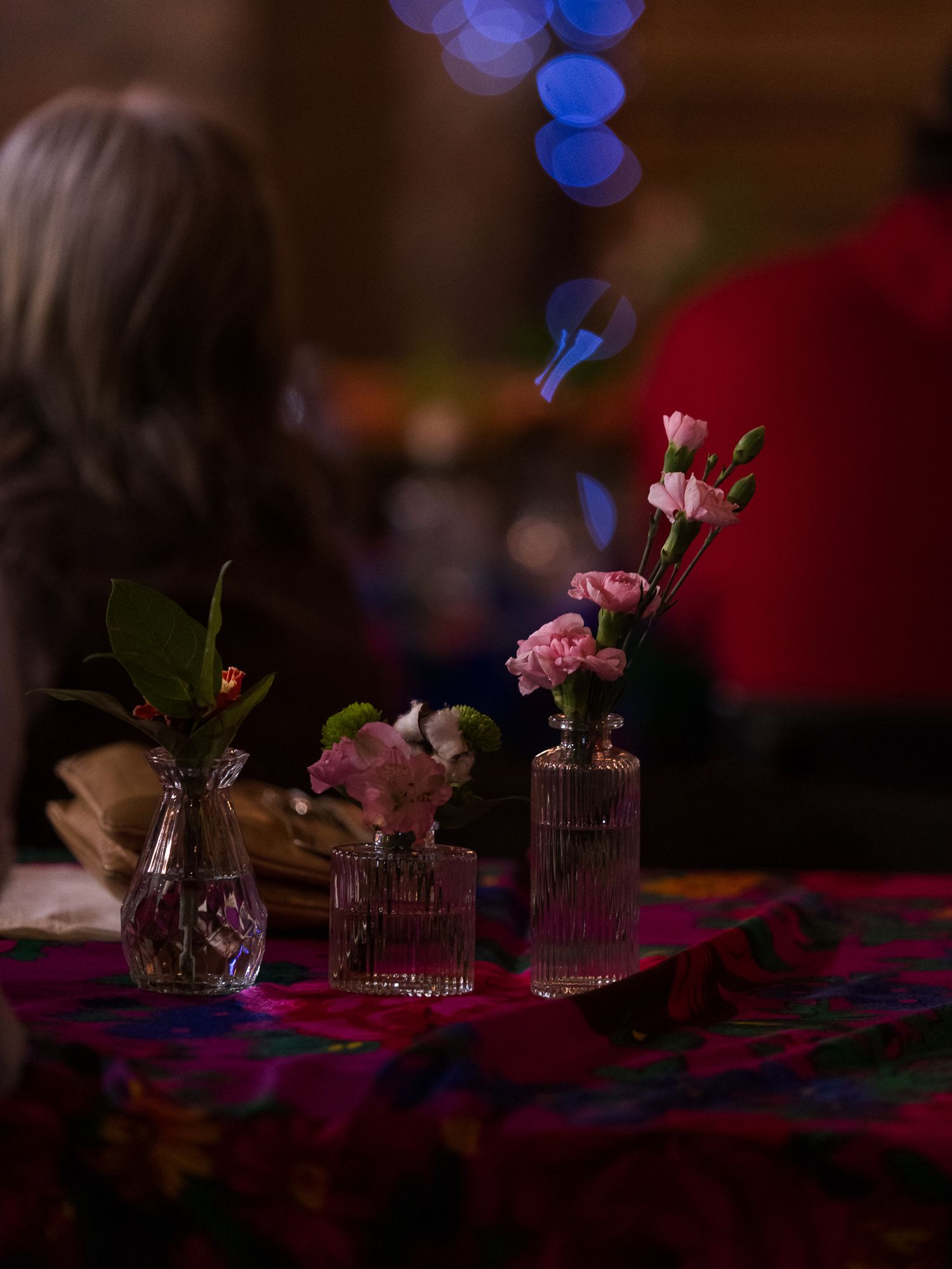 Small glass vases with pink carnations and flowers on a colorful tablecloth at an event.