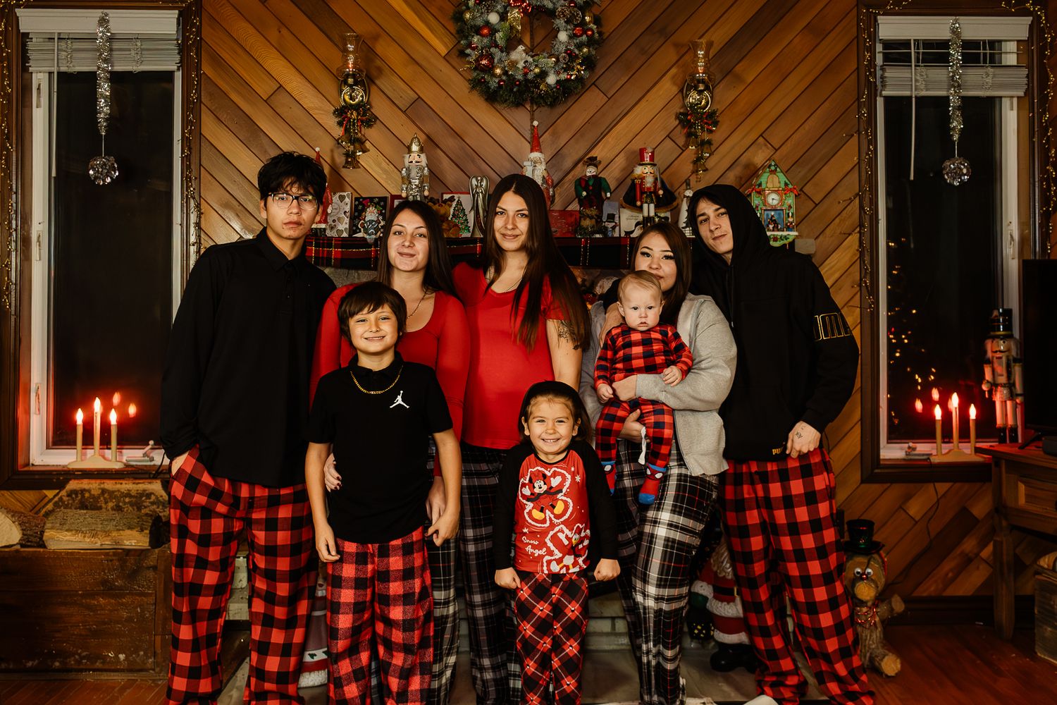 Family of seven posing in matching red plaid pajamas in front of festive Christmas decor with candles and wreath.
