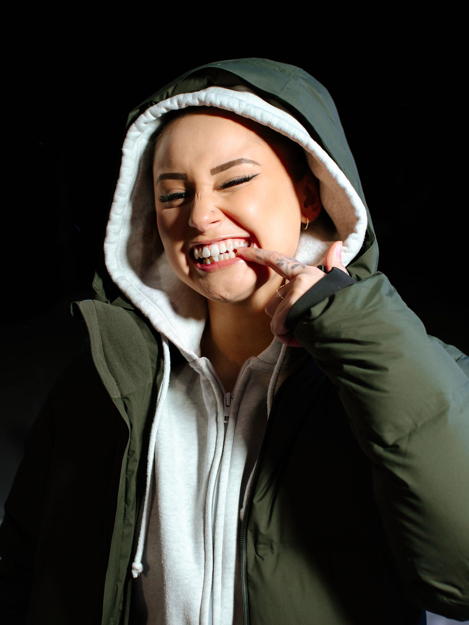 A smiling woman in a green hooded puffer jacket biting her finger against a dark background.