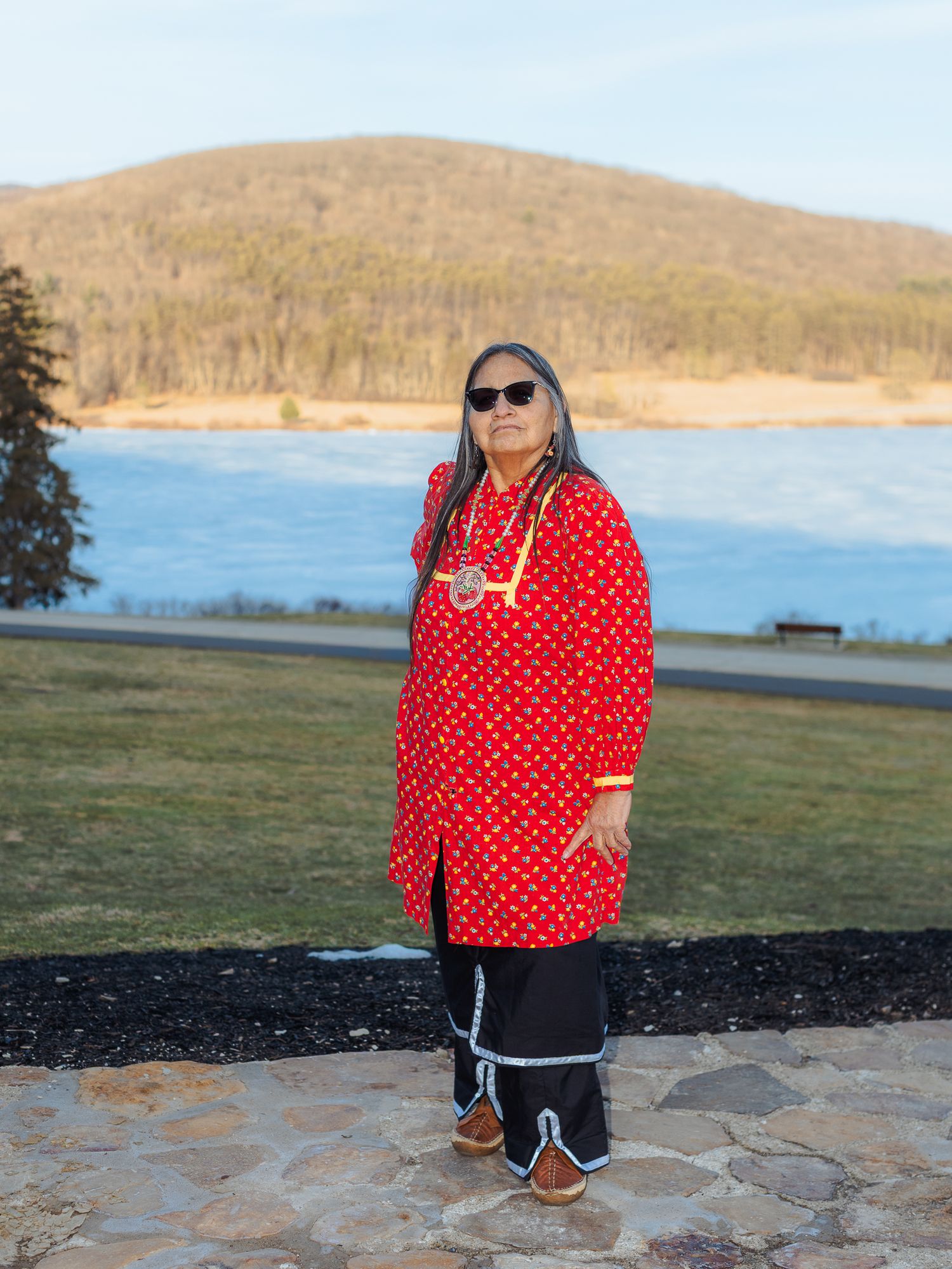 Woman in red polka dot traditional top and black pants stands before a serene lake and tree-covered mountain.