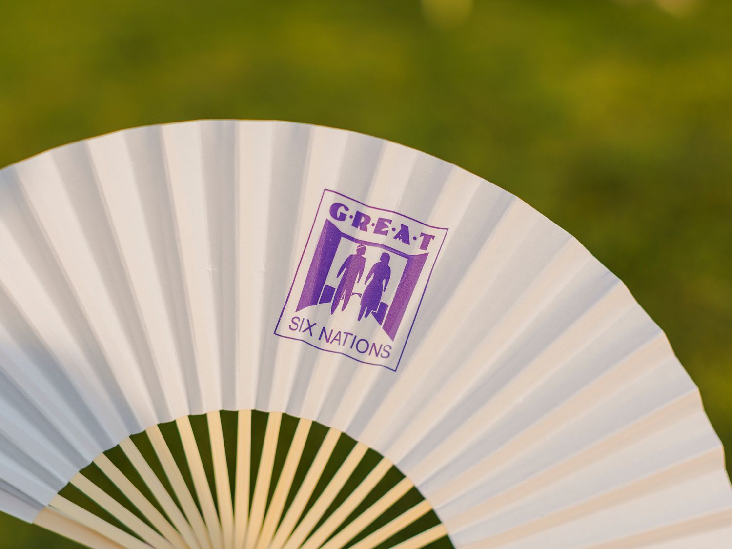 White folding fan featuring a purple Great Six Nations logo, set against a blurred green background.