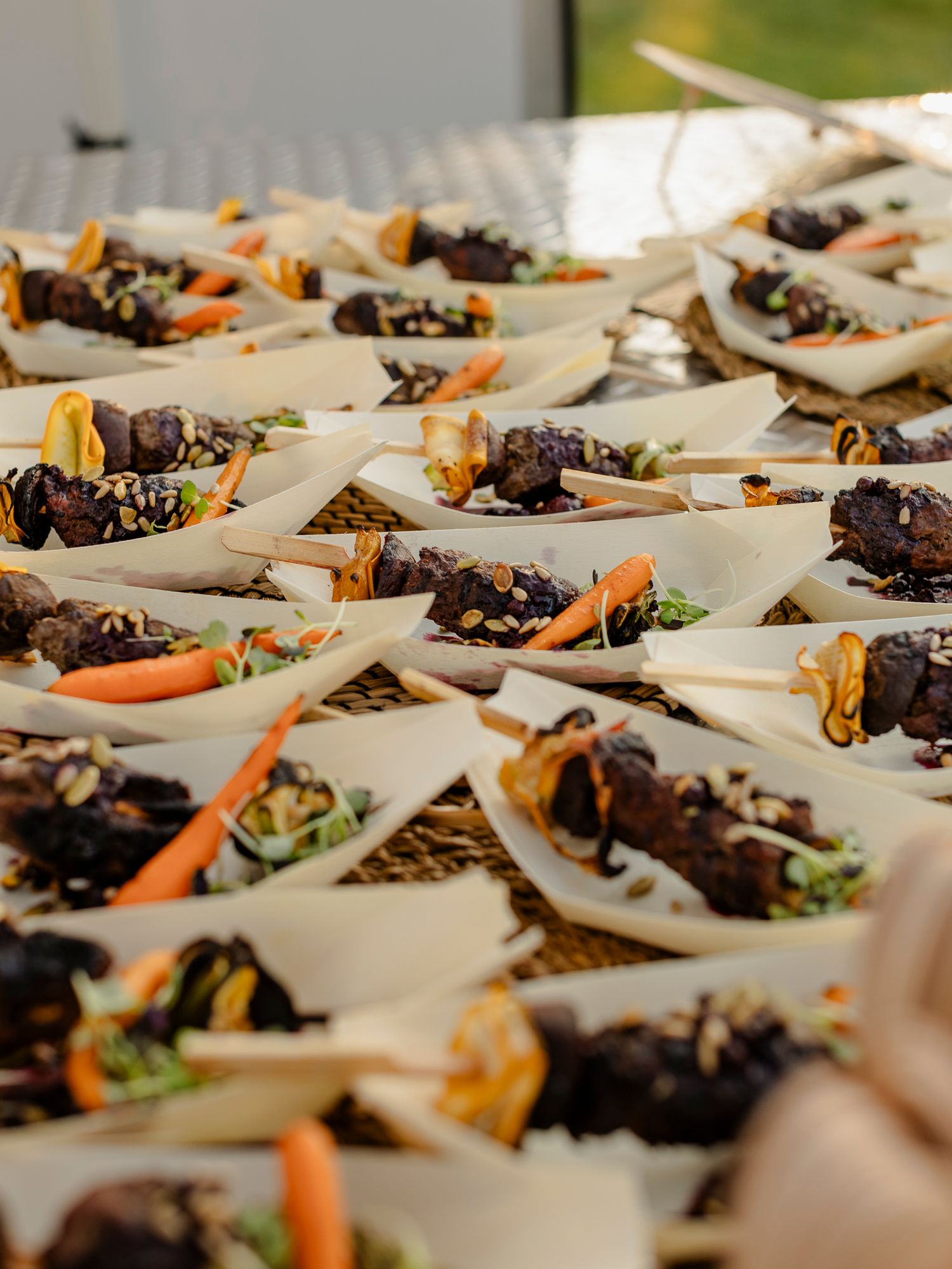 Elegant catered appetizers in paper boats with glazed meat, carrots, and sesame seeds arranged on a buffet table.
