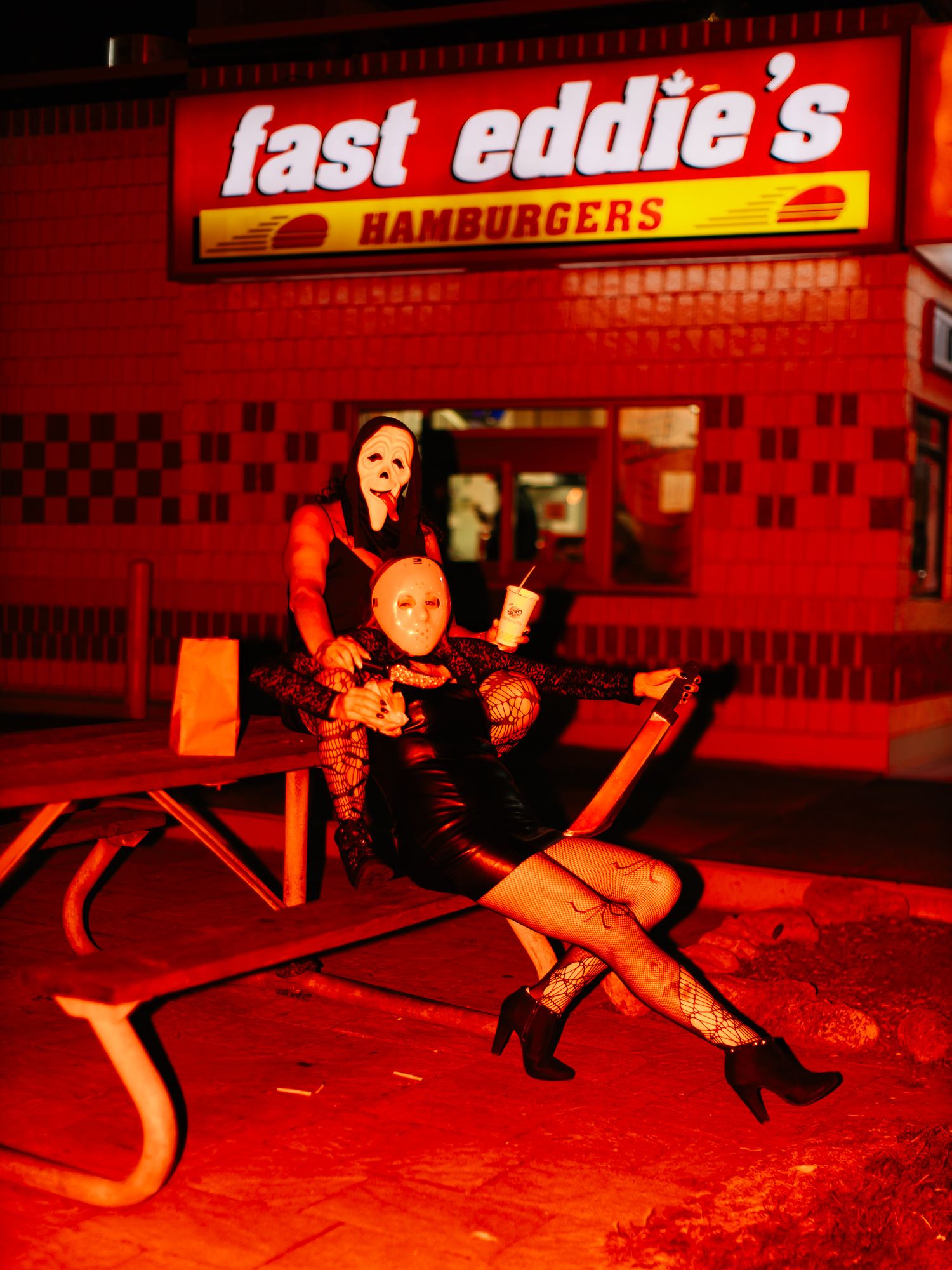 Red-illuminated figures in dark clothing posing outside Fast Eddie's Hamburgers at night.