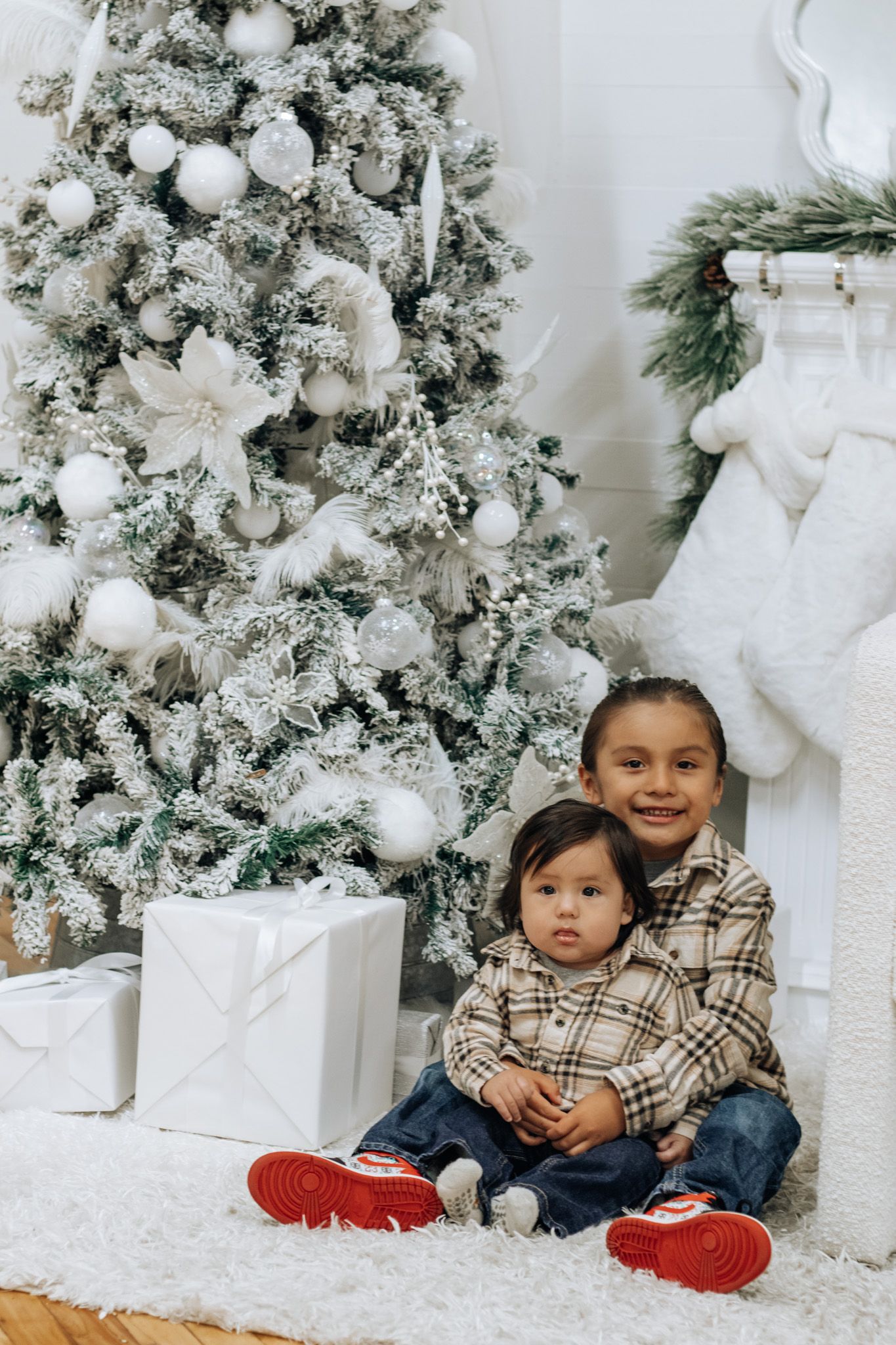Children sitting together by decorated white Christmas trees during holiday photo session.