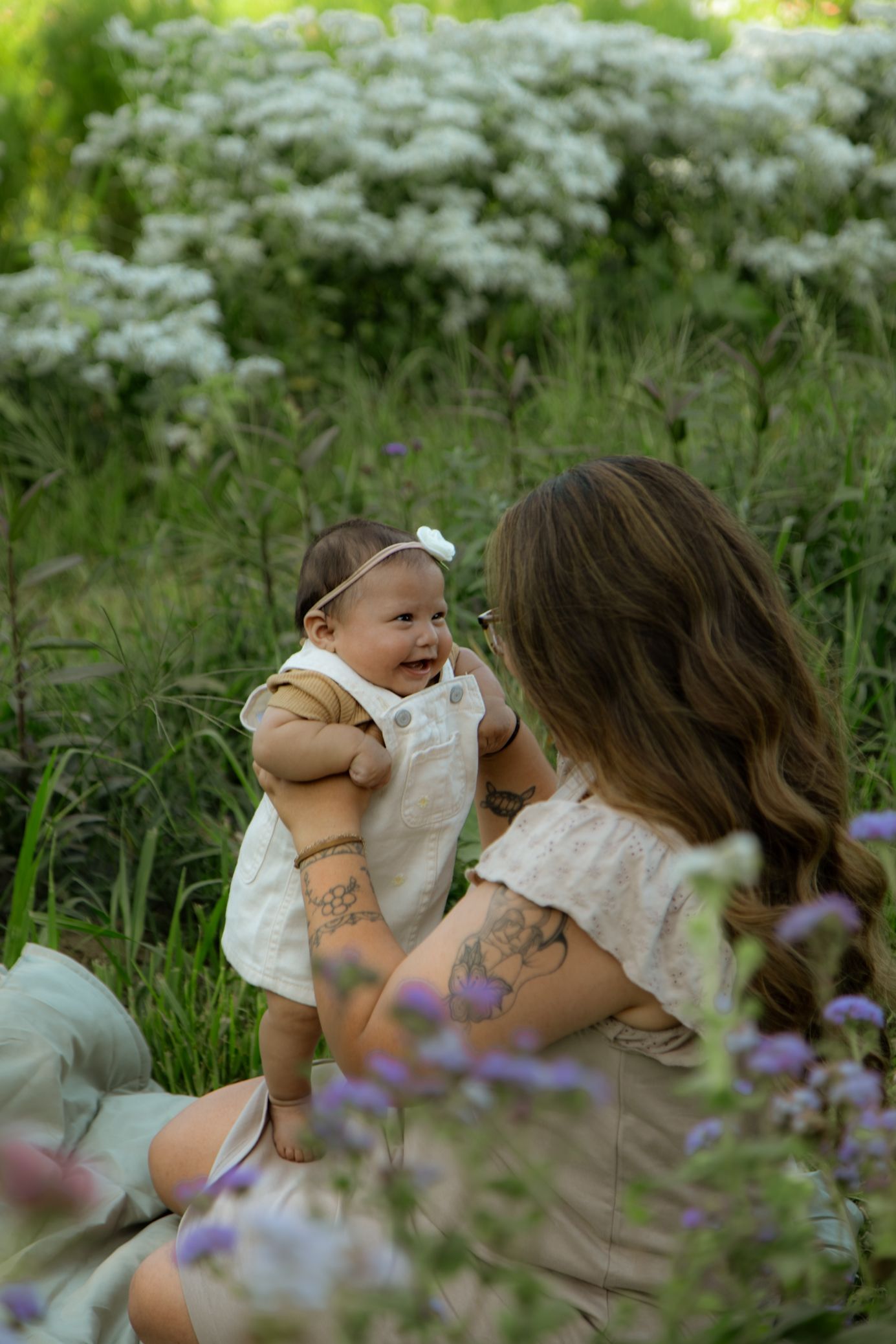 Baby in white overalls smiles while playing outdoors among blooming white flowers.