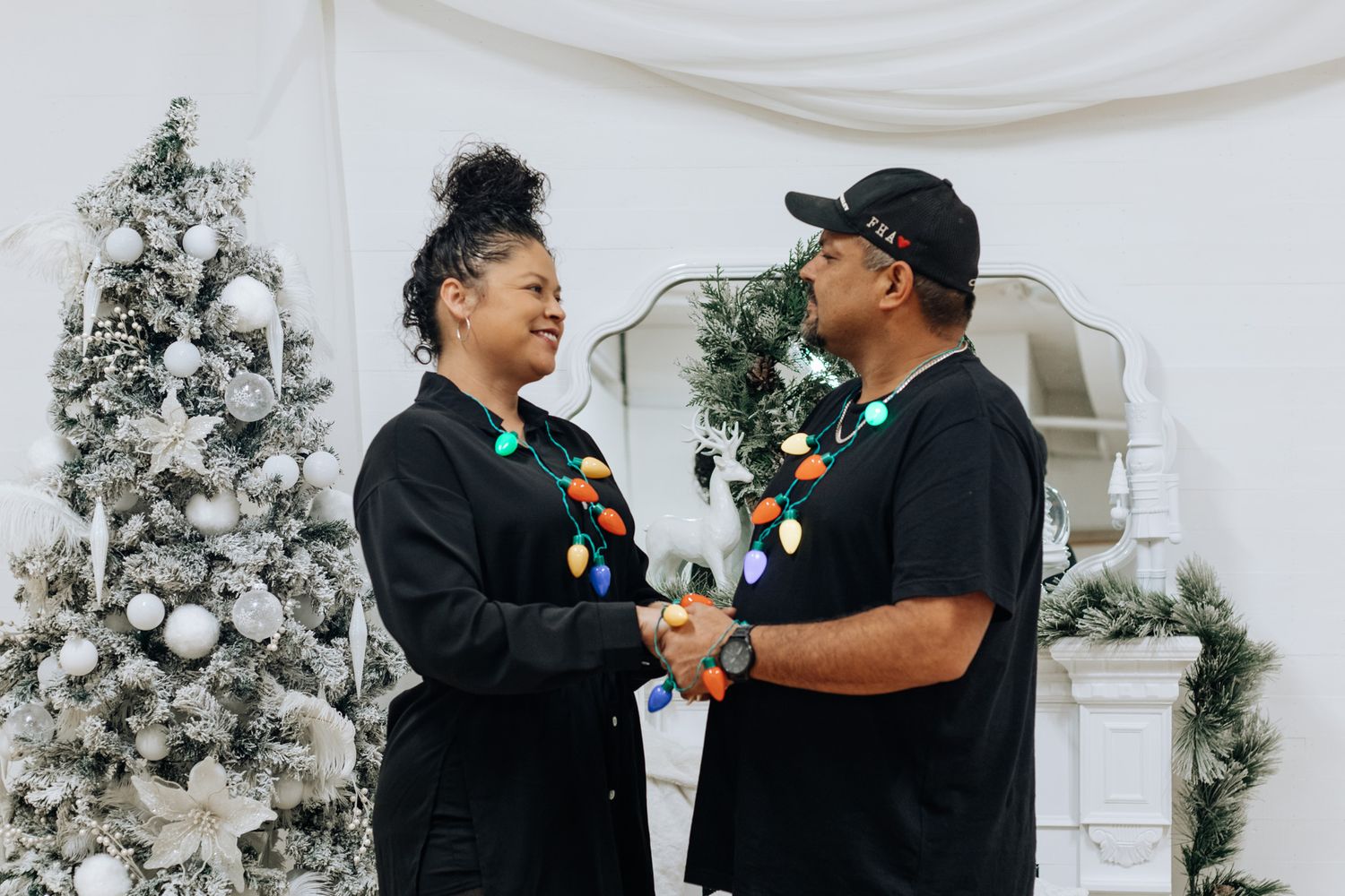A couple wearing matching black outfits shares a joyful moment by a decorated Christmas fireplace.