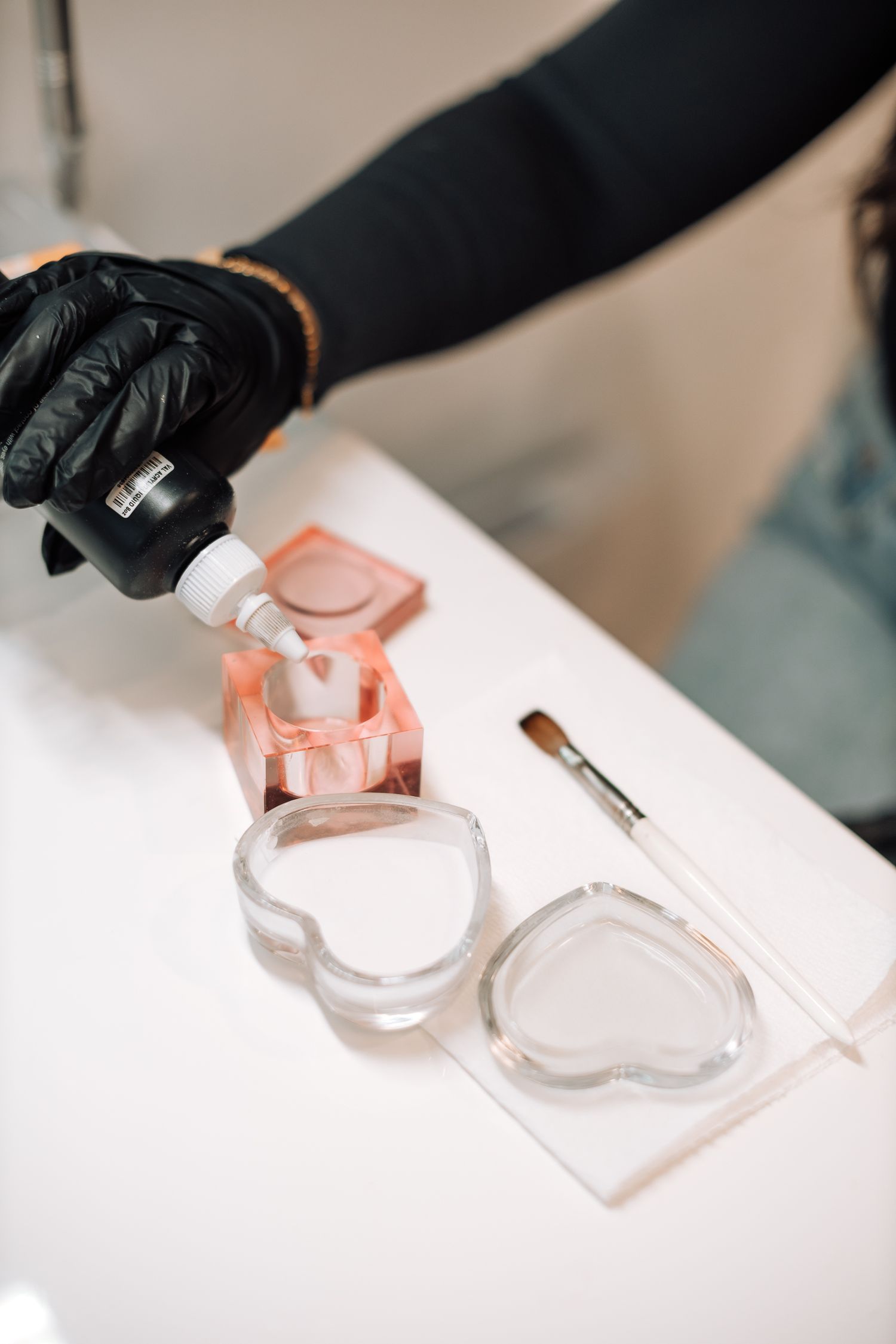 Hands wearing black gloves working with clear nail polish containers on a white surface.