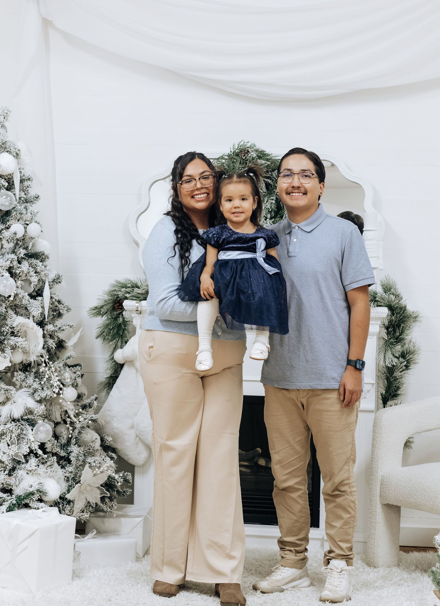Family group photo in coordinated outfits against a white Christmas backdrop with decorated trees.