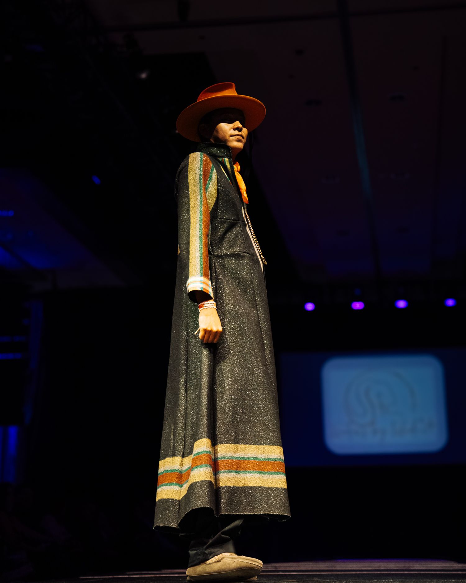 Fashion show model walking runway in traditional gray cape with orange stripe detail.
