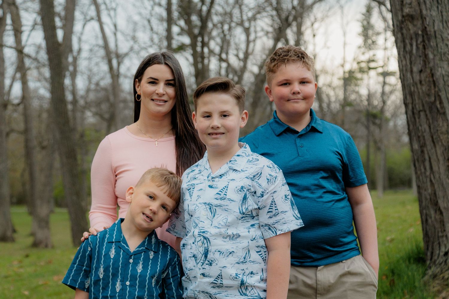 Woman in pink dress posing outdoors with three boys in blue shirts, surrounded by trees in a park setting.