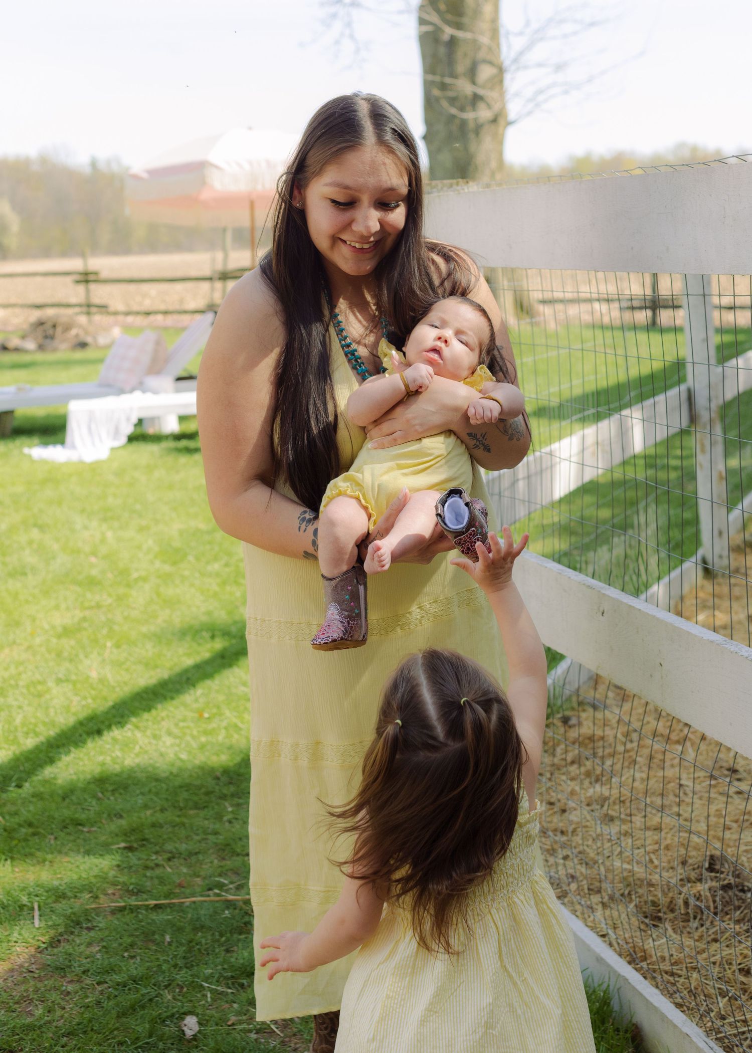 Woman in yellow dress holds baby while toddler reaches up outdoors near white fence on sunny day.