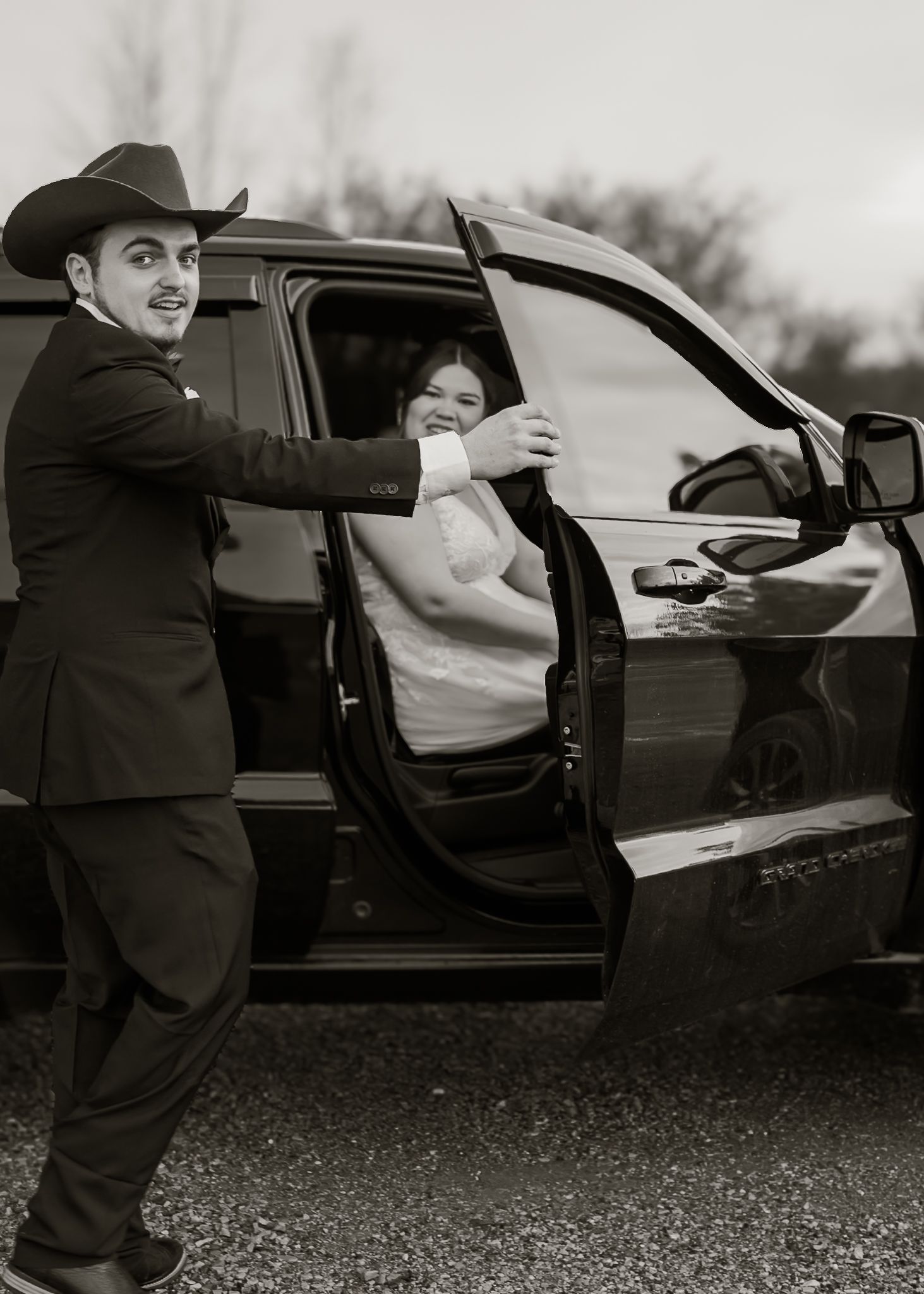 Groom in black cowboy hat opens truck door for smiling bride in white wedding dress, black and white.