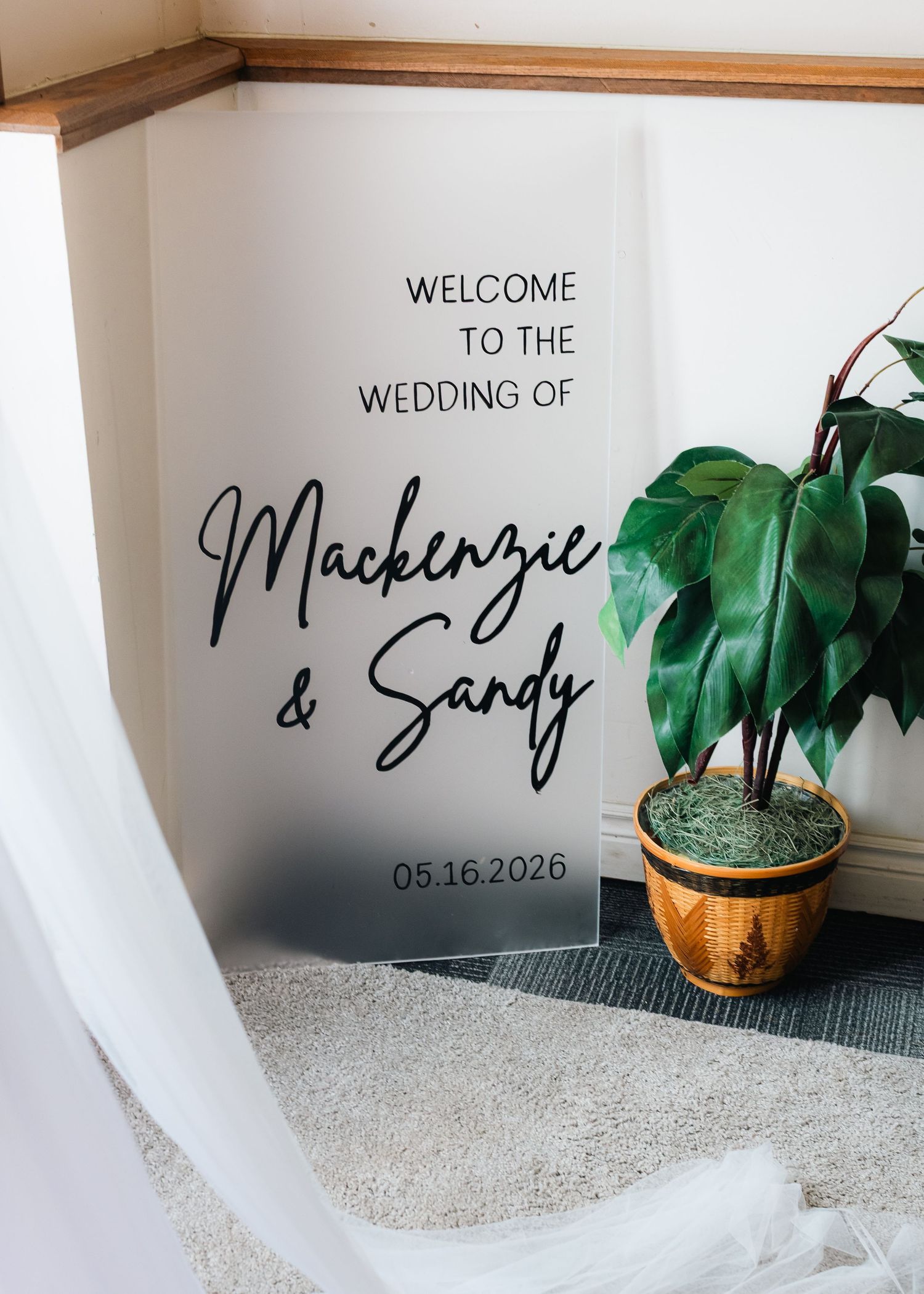 Frosted acrylic wedding welcome sign for Mackenzie and Sandy dated 05.16.2026 beside a green plant.
