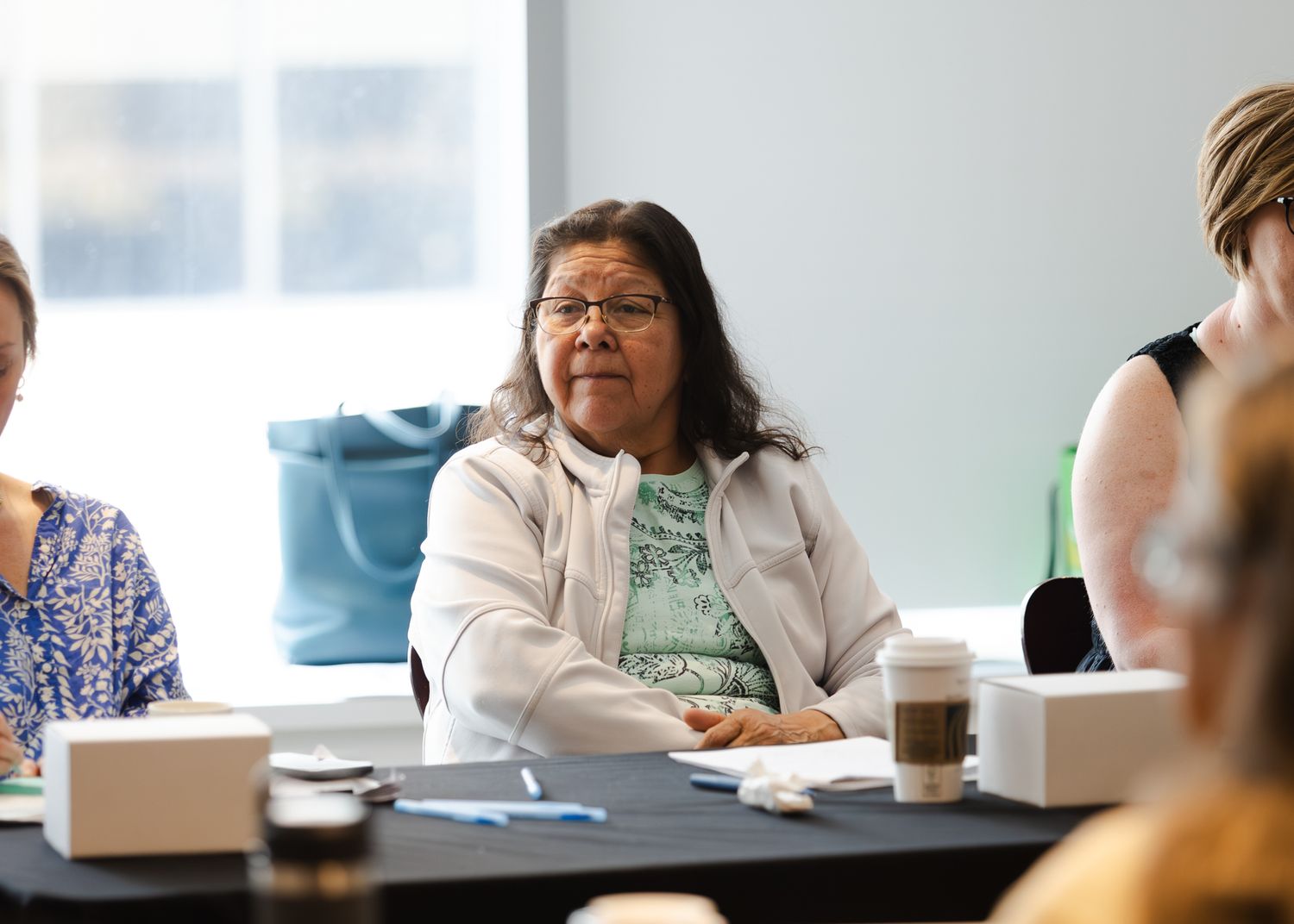 An elderly Indigenous woman with glasses sits at a conference table during a community meeting.
