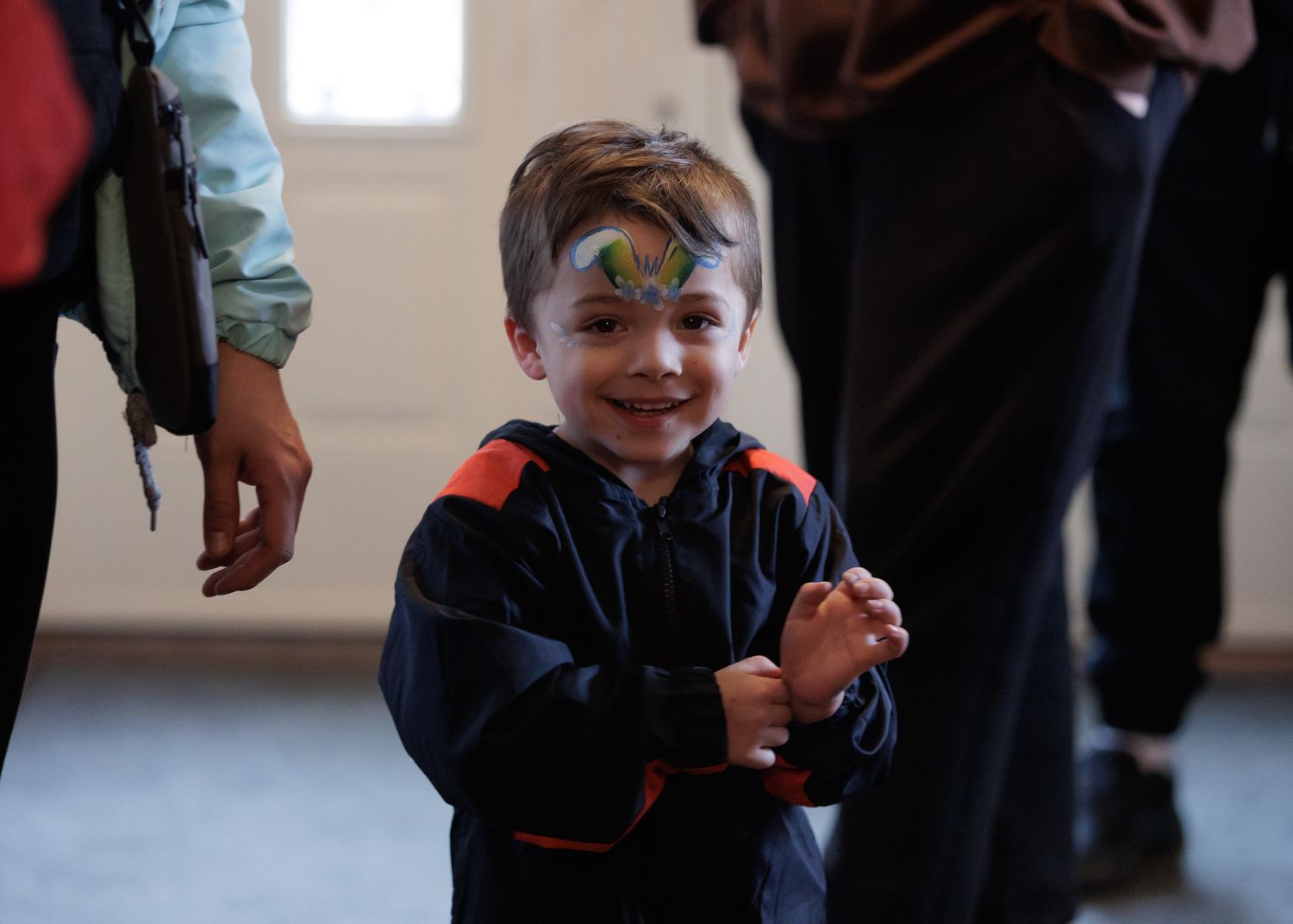 Smiling young boy with face paint clapping hands at an indoor event, wearing a dark blue jacket.