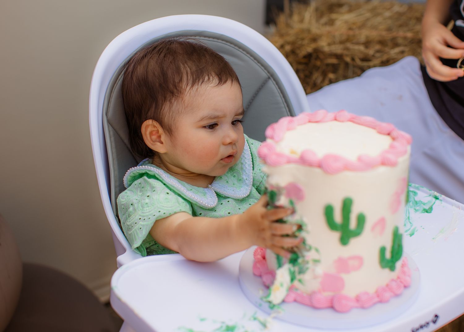 A baby girl in a green outfit touches her first birthday cake decorated with pink flowers and cactus designs.