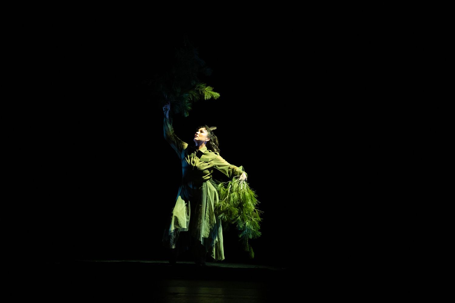 Performer in green costume dancing dramatically under spotlight on dark stage.