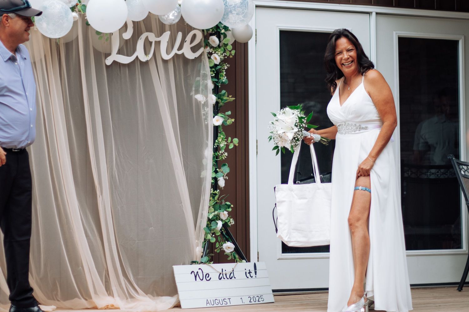Radiant bride in white gown holds bouquet near Love balloon backdrop at outdoor wedding.