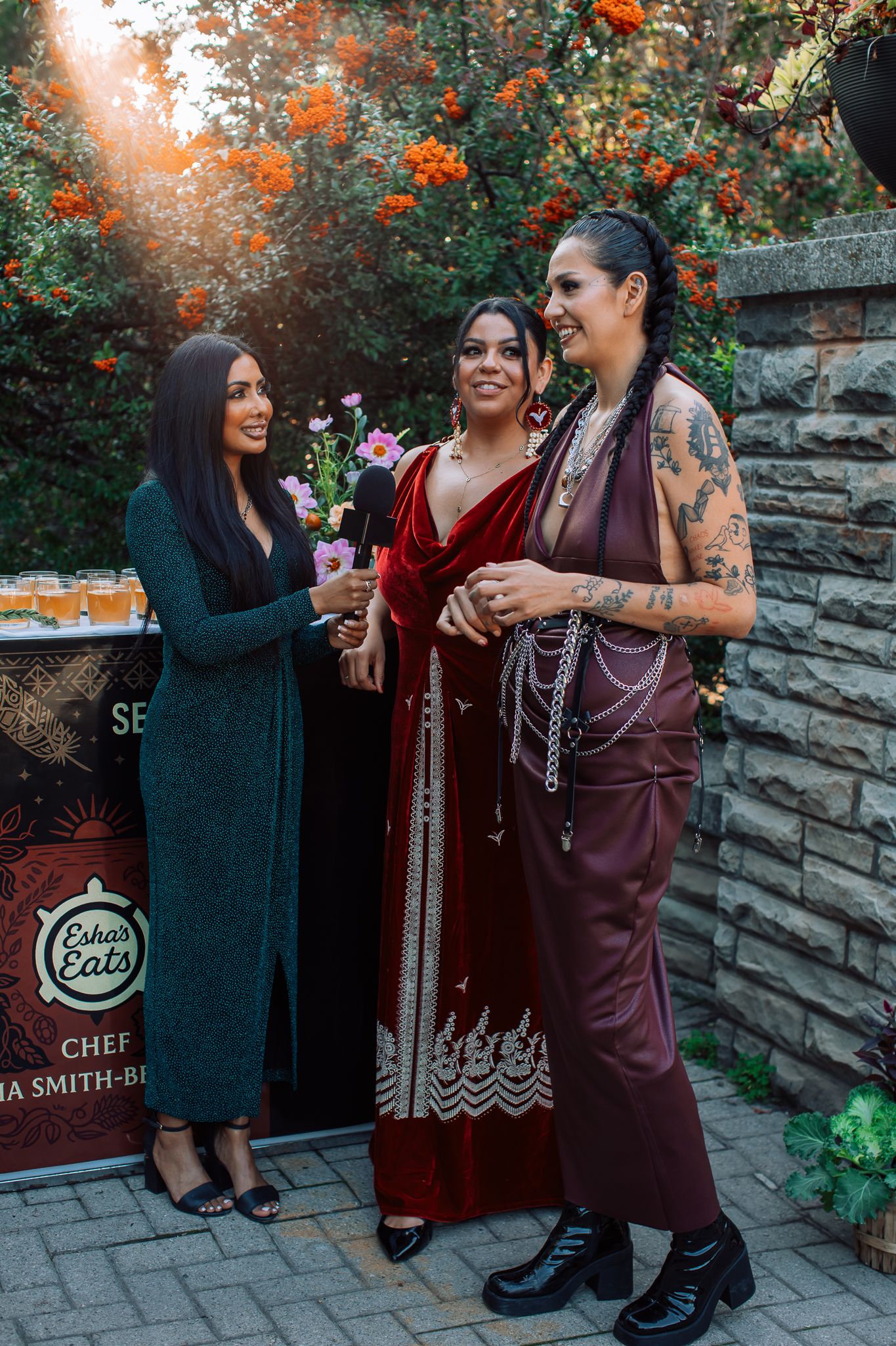 Three women in elegant outfits chat and smile at an outdoor evening event with floral backdrop.
